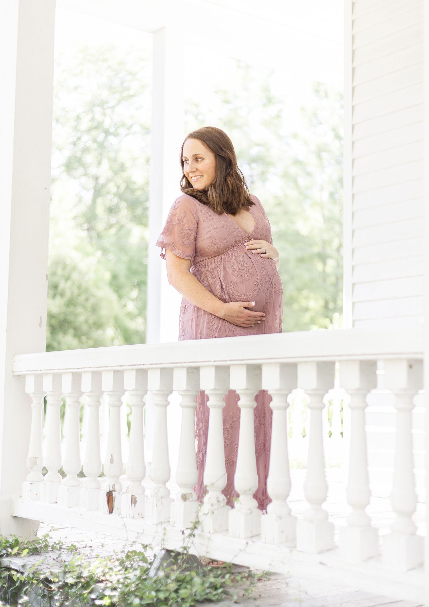 Pregnant woman in pink lace dress cradling baby bump on white balcony porch surrounded by lush greenery.