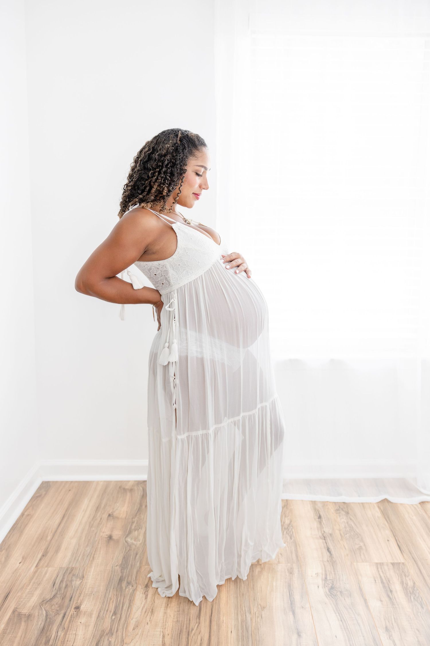 Pregnant woman in sheer white maxi dress cradling baby bump, standing in bright white studio with wood floors.