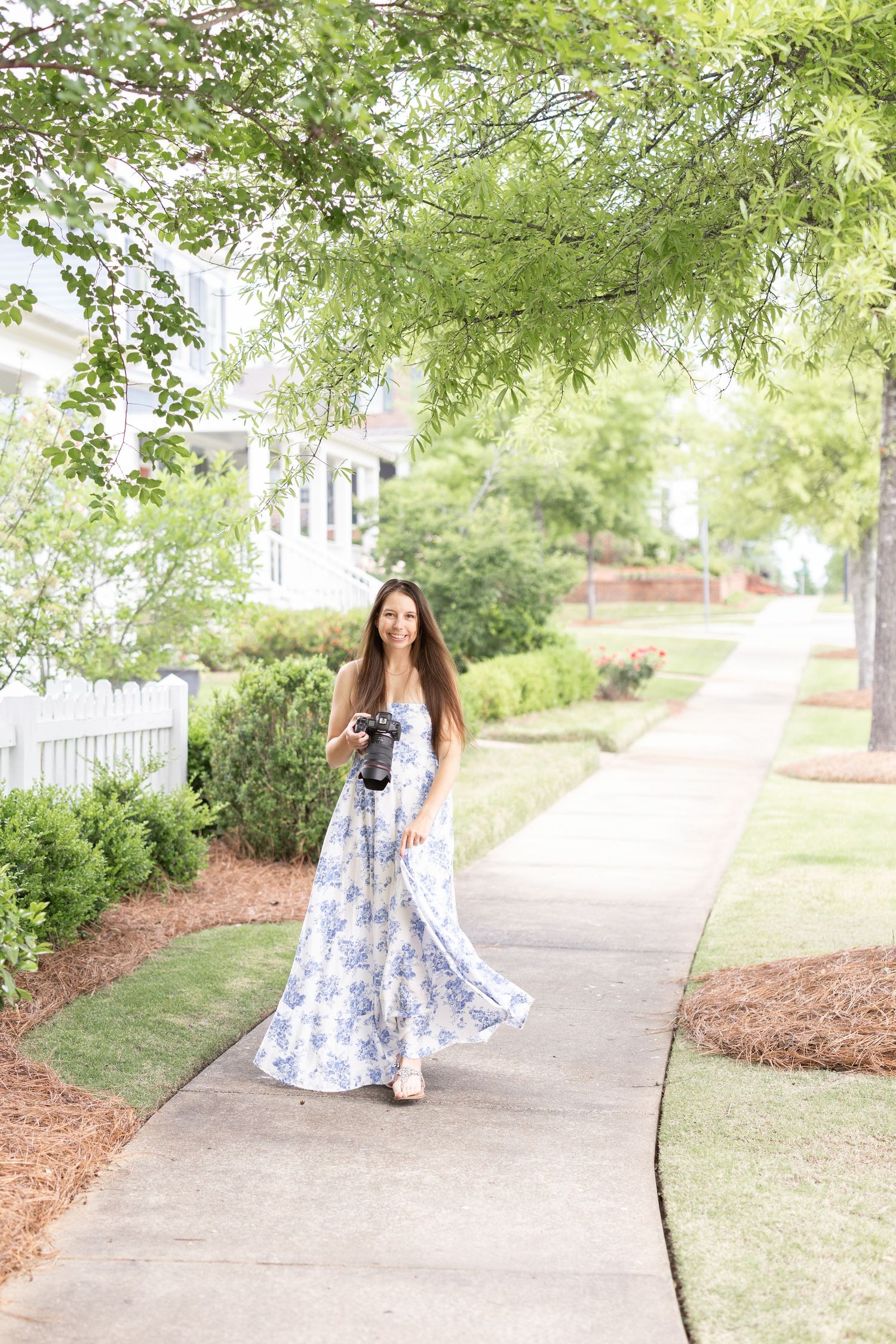 Woman in blue floral maxi dress holding camera walks on suburban sidewalk surrounded by lush green trees.
