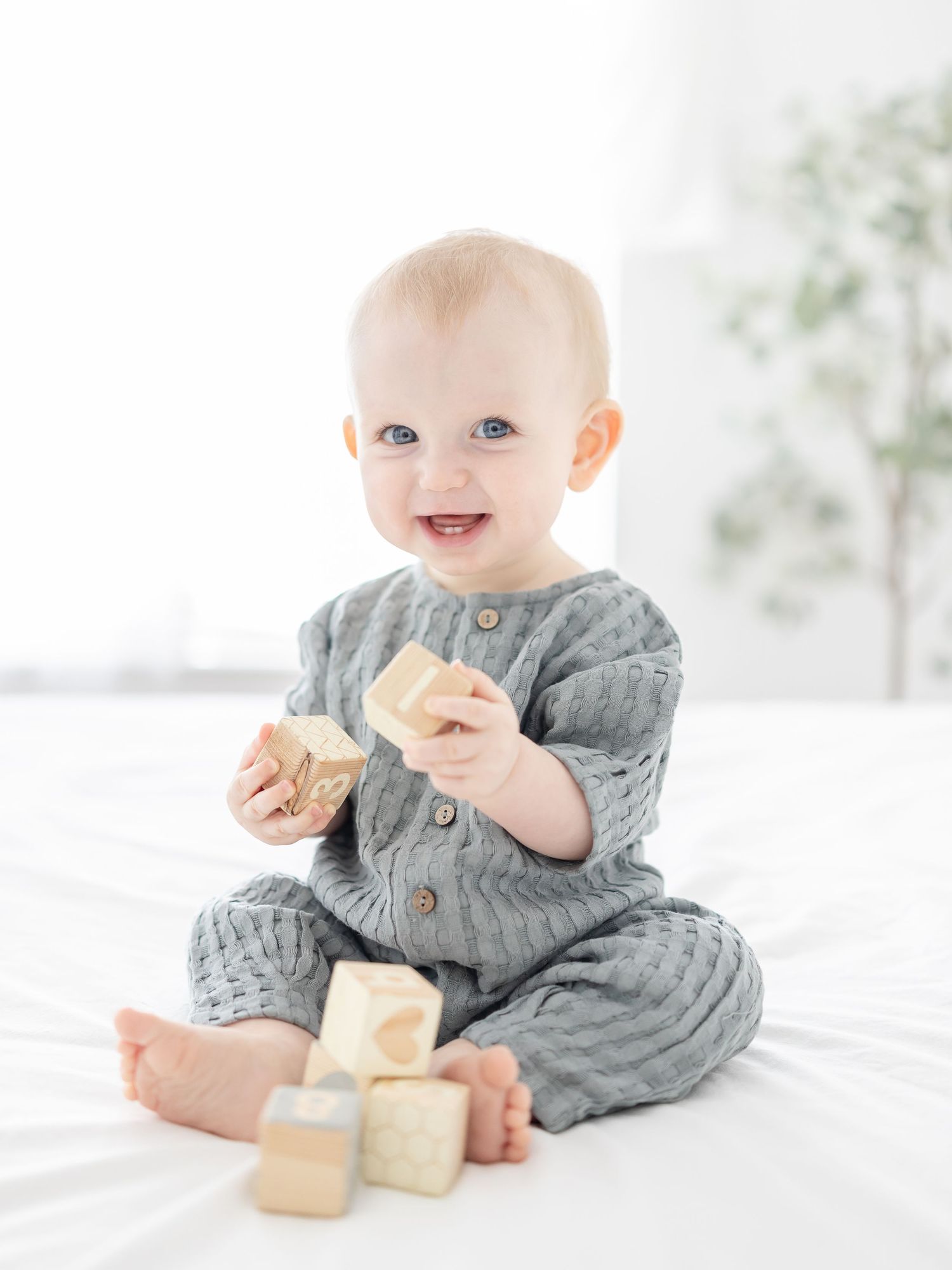Baby in blue jumper sits on a white bed playing with wooden blocks at his nine month milestone session by Columbus, GA baby photographer.