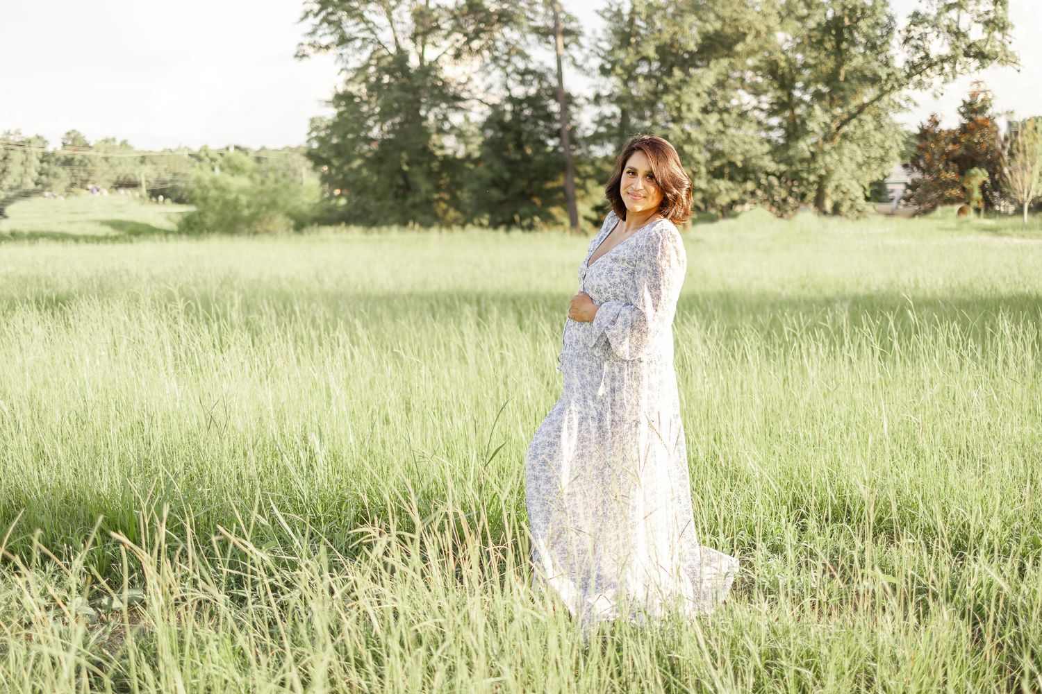 A floral sundress adorned figure twirls gracefully in a sun-drenched meadow with tall grass and trees in the background.