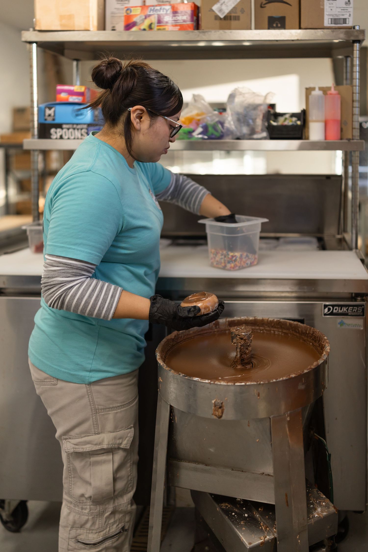 Worker in turquoise shirt operates industrial chocolate tempering and mixing equipment in commercial kitchen setting.