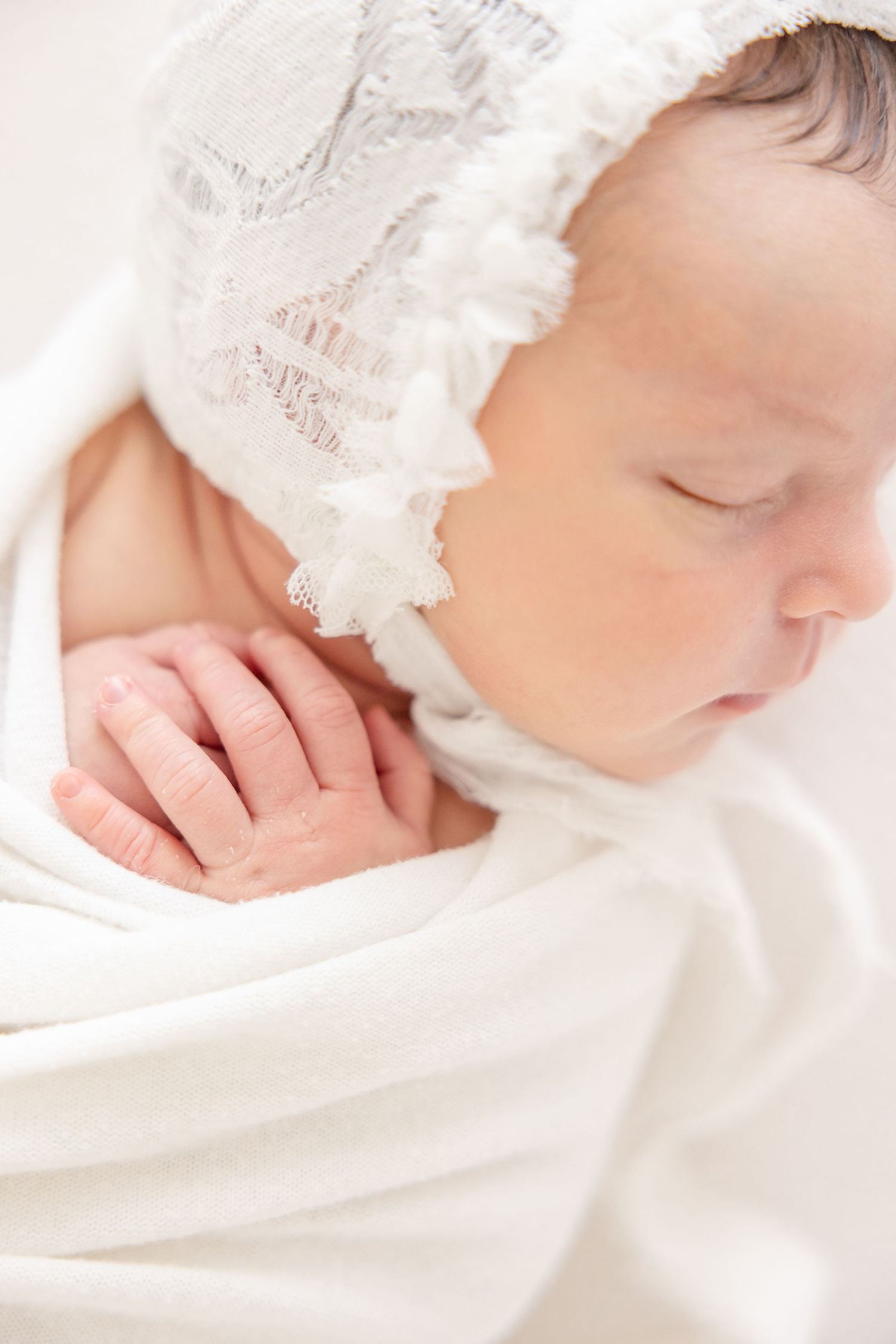 Sleeping newborn baby wrapped in white swaddle wearing a delicate lace bonnet, peaceful close-up portrait.