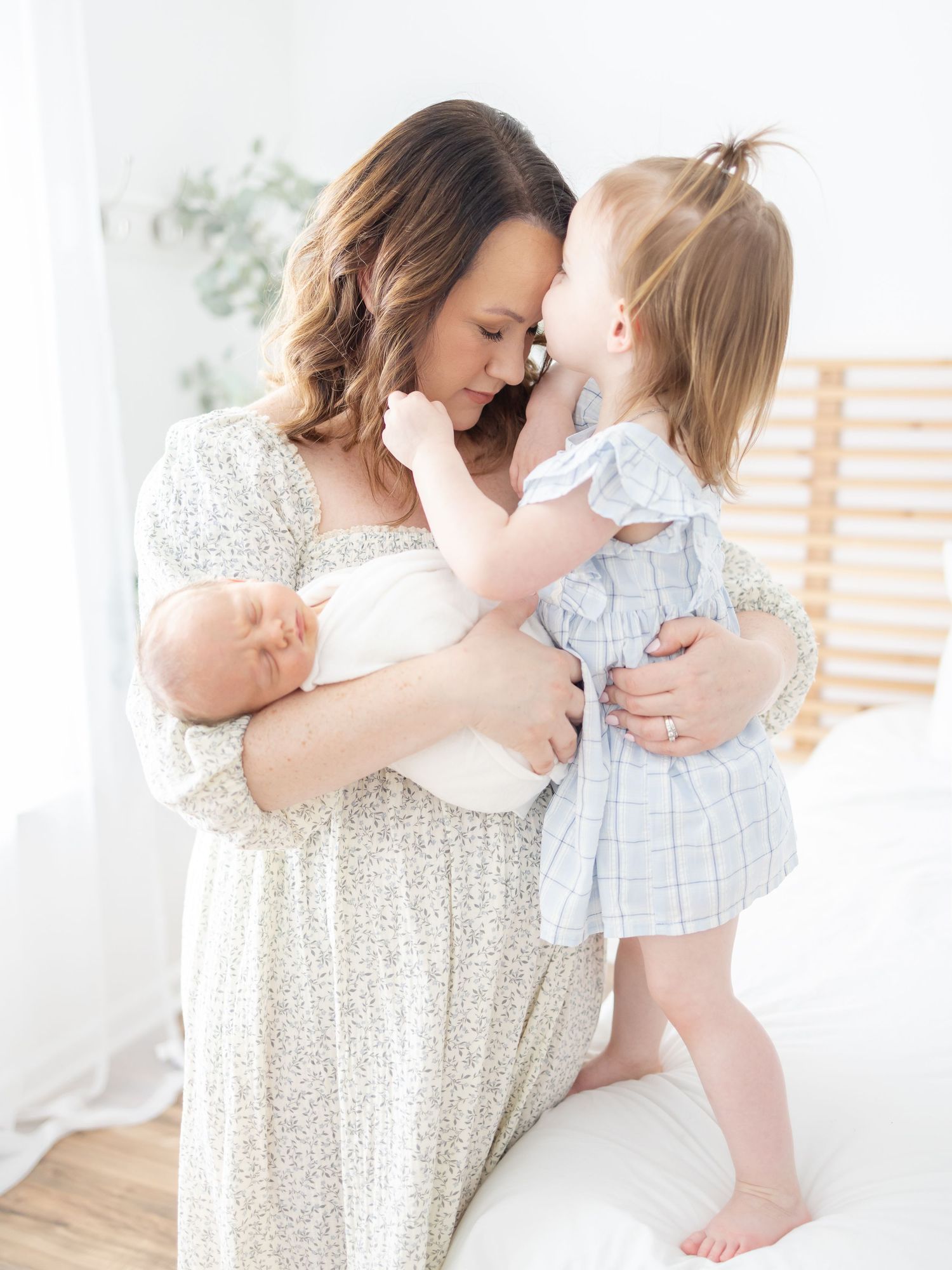 A mother in a floral dress holds a newborn while her toddler daughter in a blue dress touches foreheads with her.