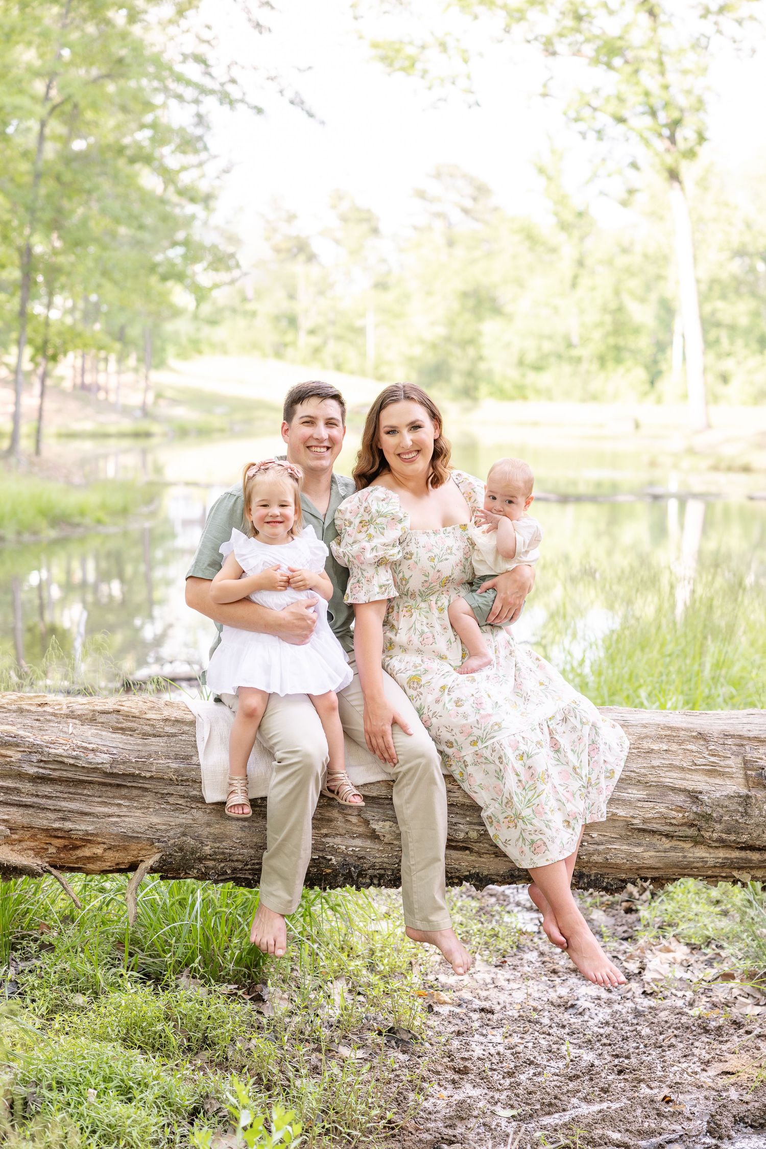 Family of four sitting on a log by a pond, parents holding two young children, smiling outdoors in soft natural light.