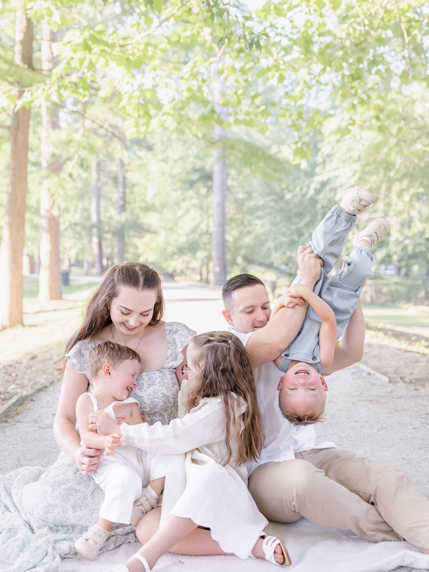 A joyful family of five sits outdoors on a blanket, with dad playfully holding a toddler upside down among tall trees.
