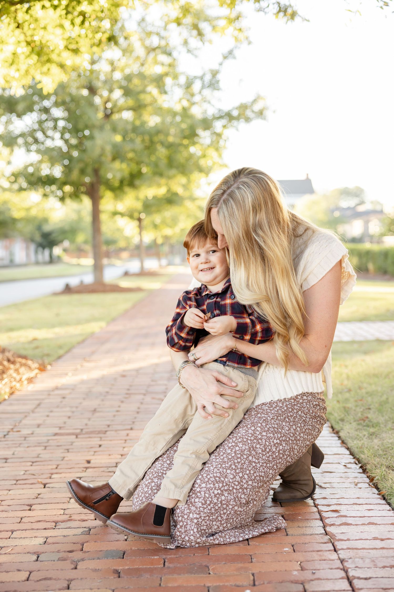 A heartwarming moment with mom kneeling on a brick pathway snuggling her son surrounded by autumn trees during their fall family photos.