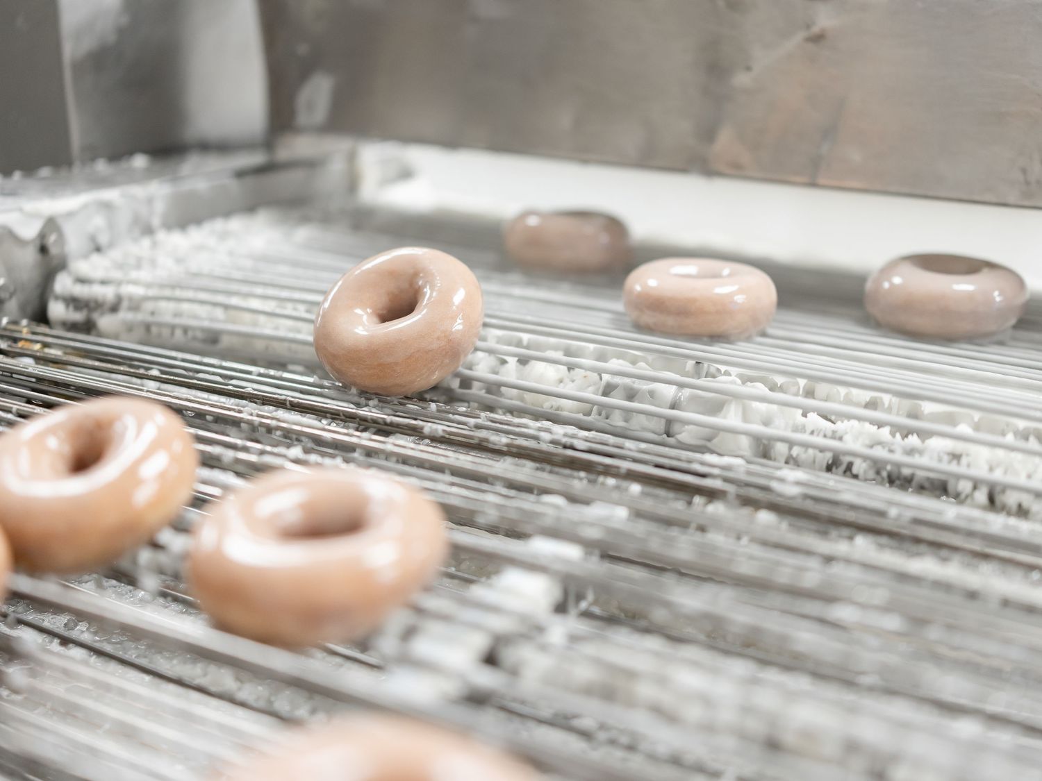 Fresh glazed donuts move along a metal conveyor belt in a commercial bakery production line.