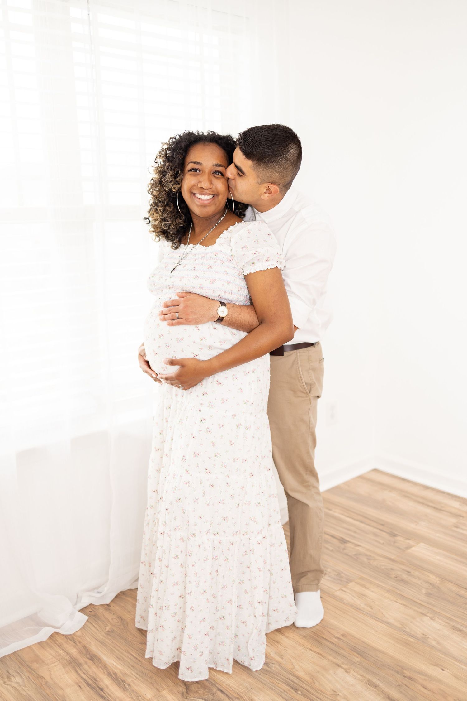 A loving couple embraces in a maternity photo shoot, wearing white and khaki against a bright minimal background.