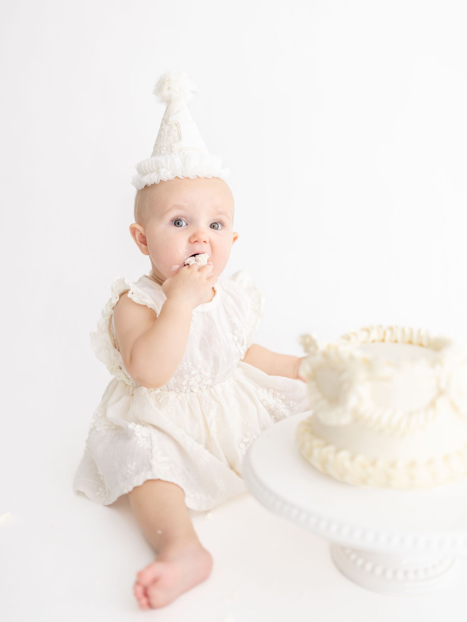 Baby girl in white dress and party hat smashes first birthday cake in a bright white studio photo session.