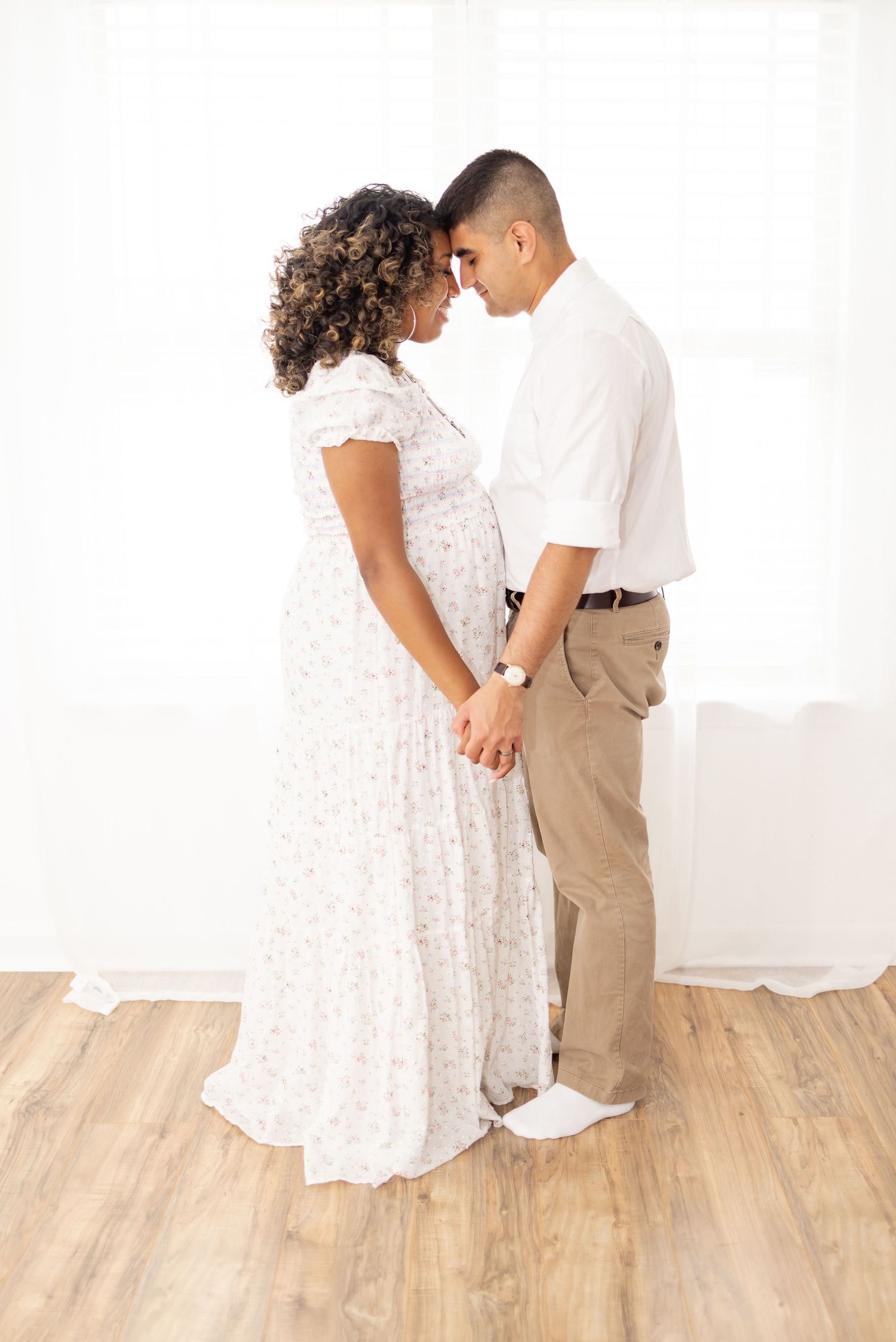 Two people in romantic pose wearing white and khaki standing close together on wooden floor against white backdrop.