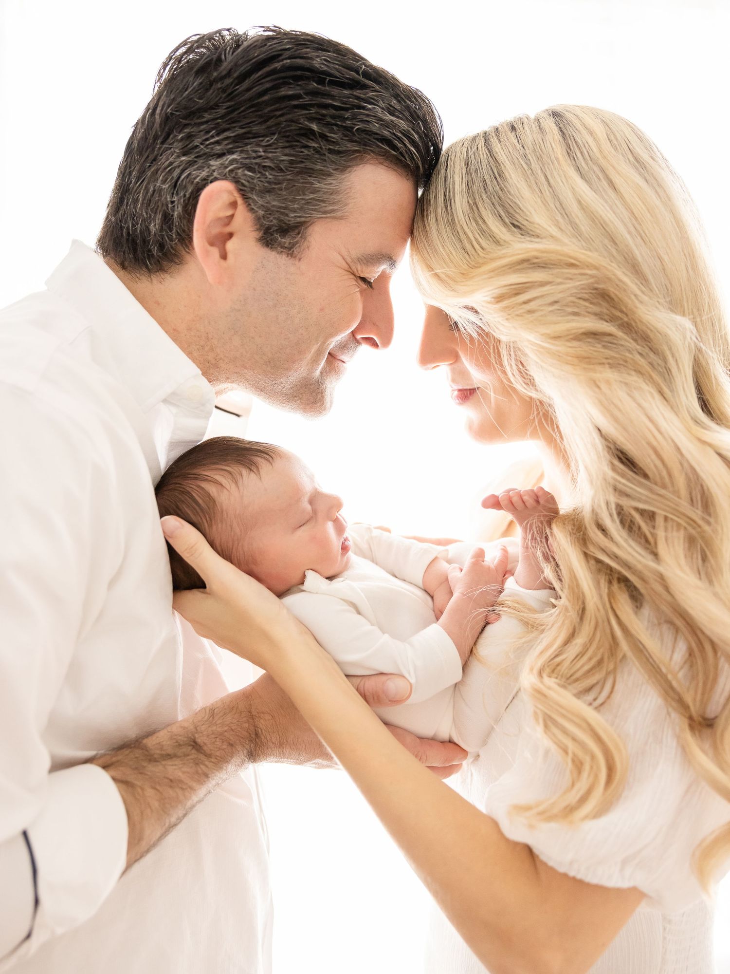 New parents dressed in white rest their heads together with their newborn daughter in between them by Columbus, GA newborn photographer.