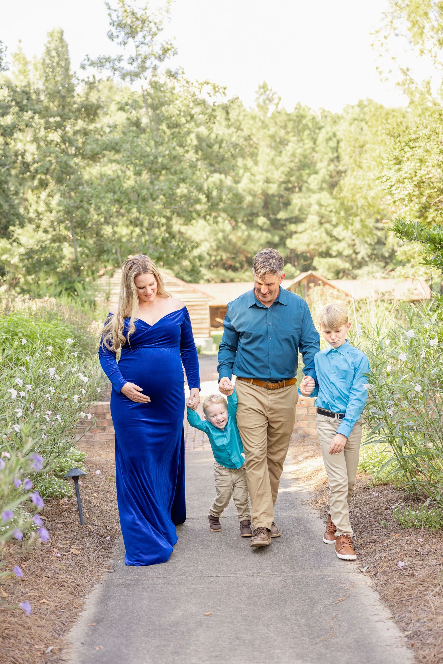 Pregnant woman in blue gown walks with husband and two boys on garden path during maternity photo session in Columbus, GA.