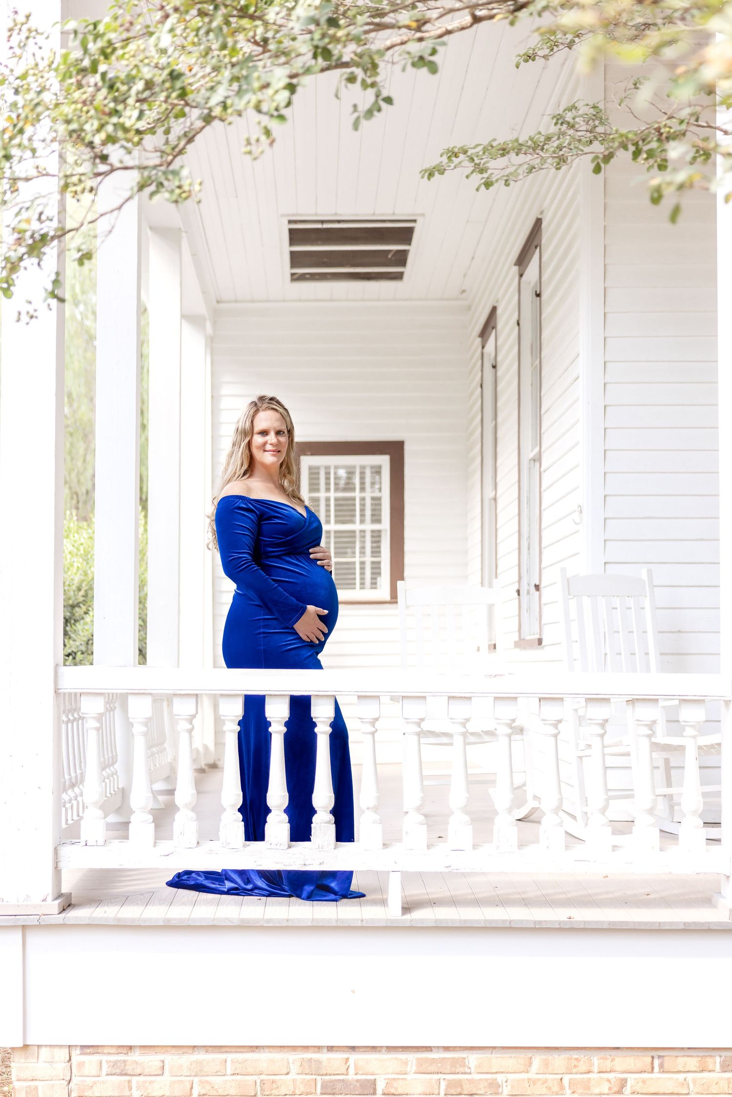 Pregnant woman in royal blue gown poses on porch of historic home during maternity photo session by Columbus, GA maternity photographer.