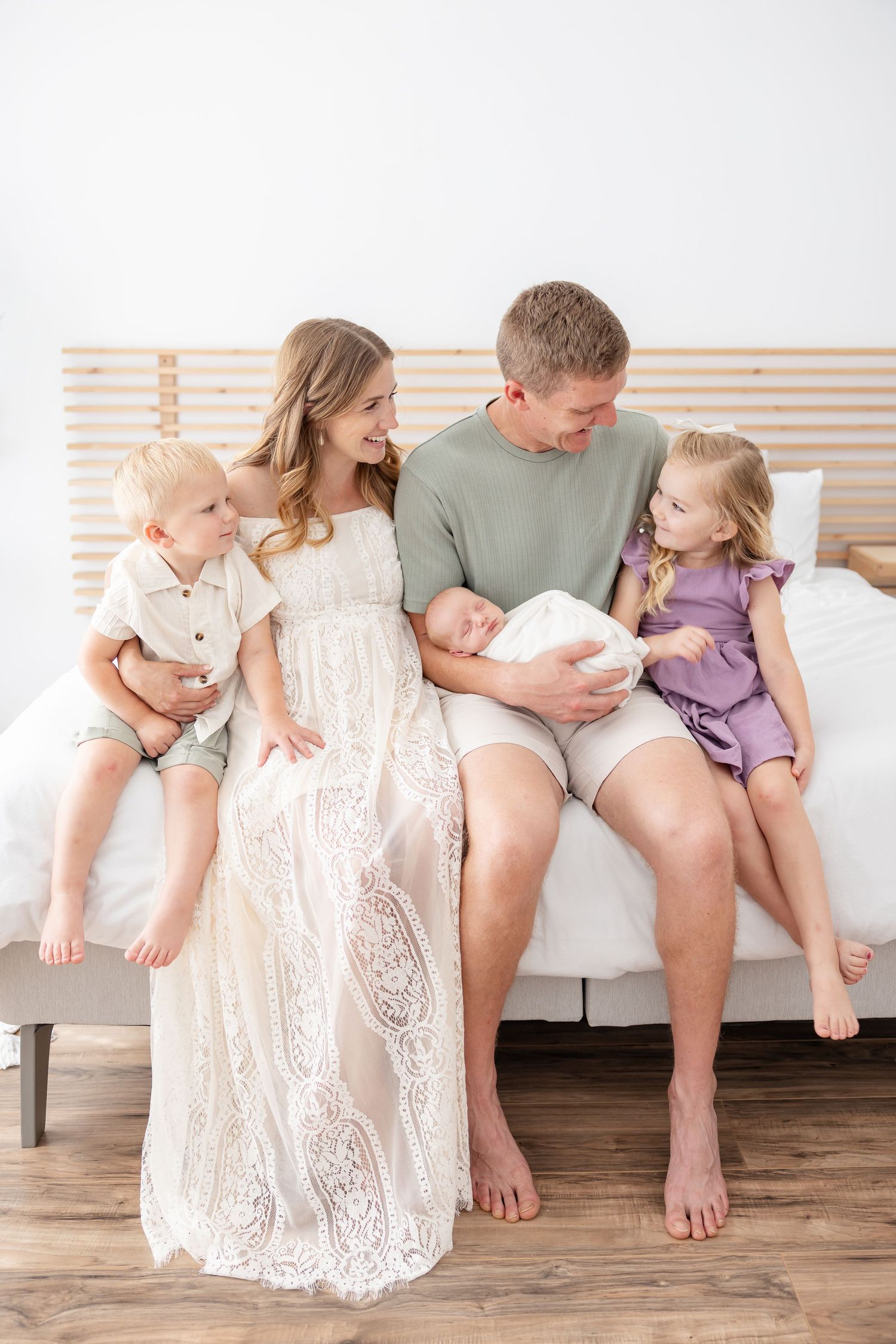 Happy family of five sitting on a bed, parents admiring their newborn baby flanked by two young children.