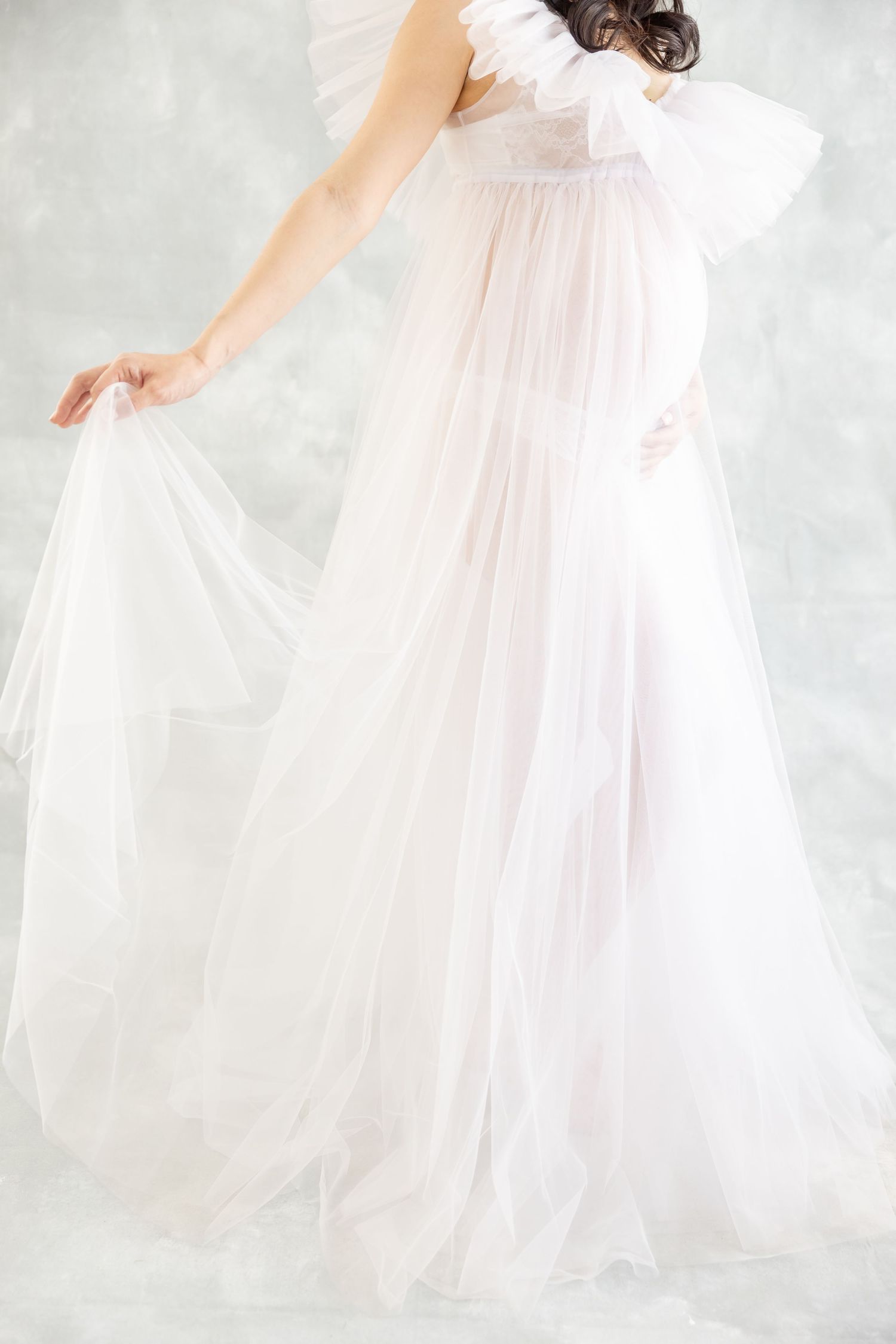 Pregnant woman in flowing white sheer tulle maternity gown posing gracefully in a soft gray studio setting.