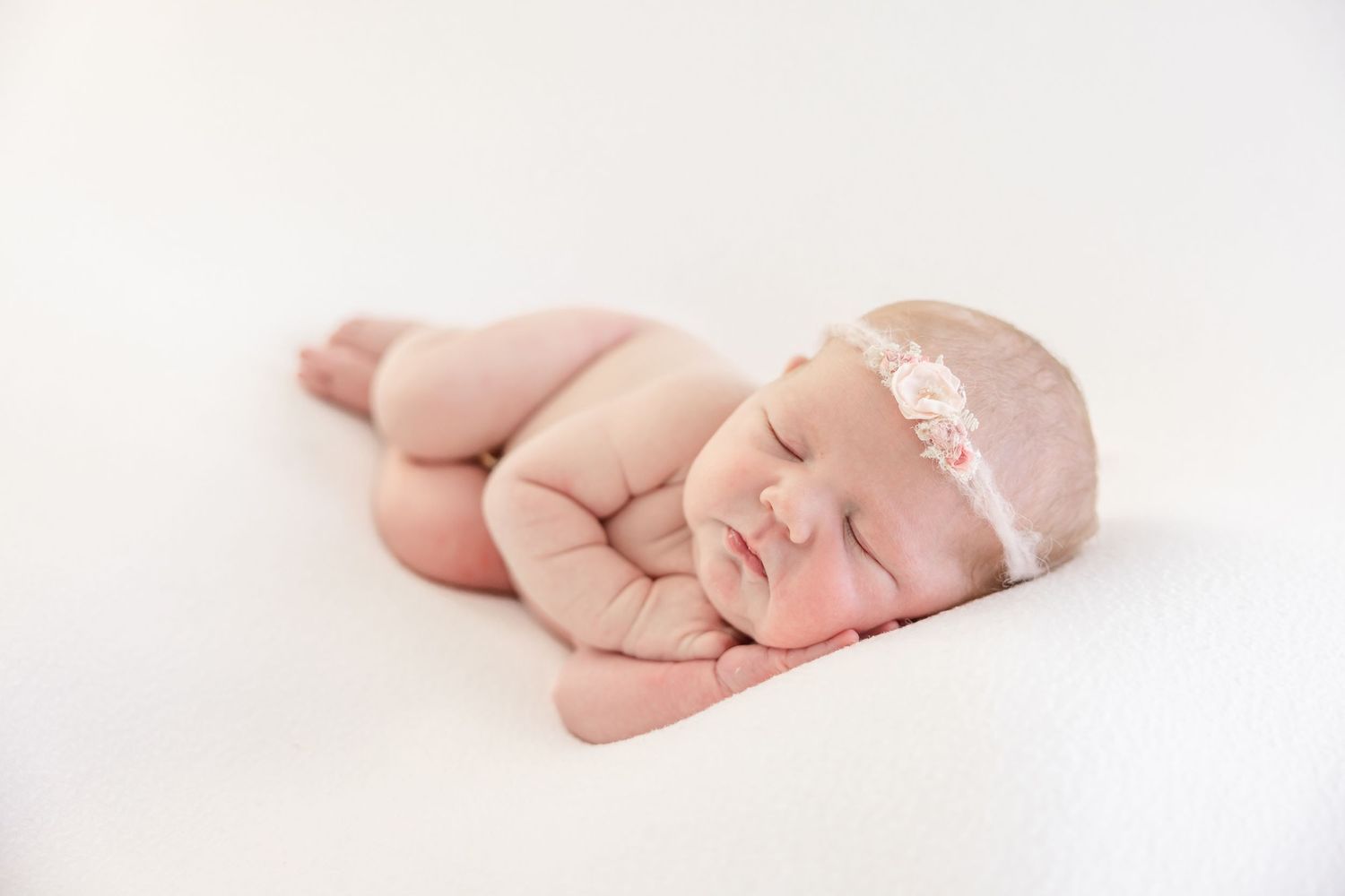 Peaceful newborn baby girl sleeping on white blanket wearing a delicate pink floral headband in a professional photo.