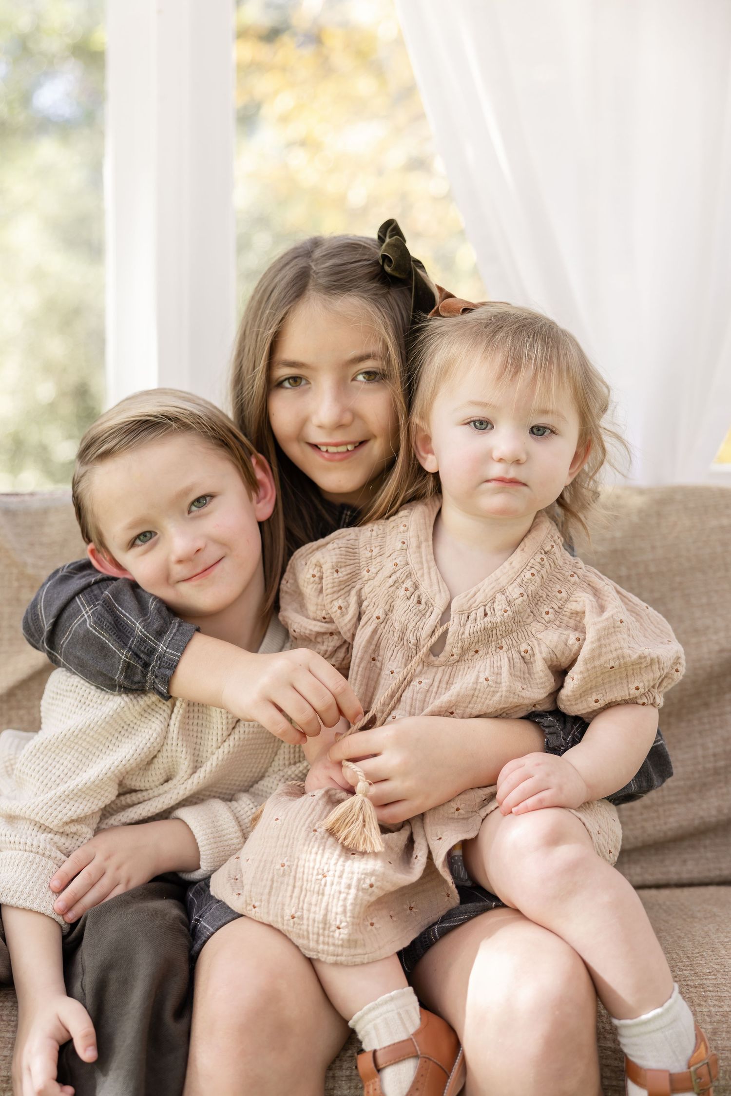 Three children sit together on a brown couch in coordinating beige outfits with natural lighting from windows behind them.
