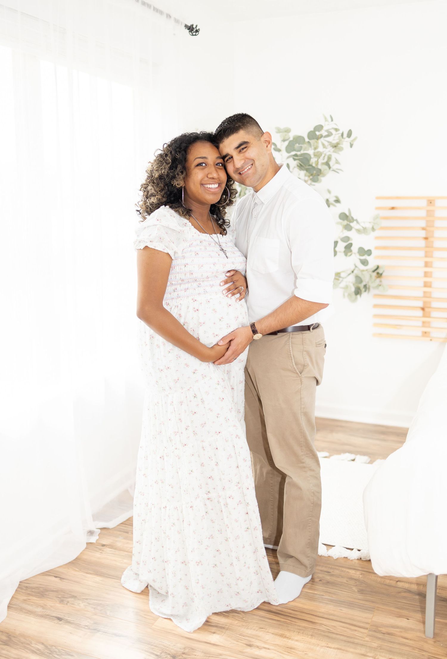 Expectant couple in white dress and khaki pants embrace lovingly in a bright minimalist room with wooden floors.
