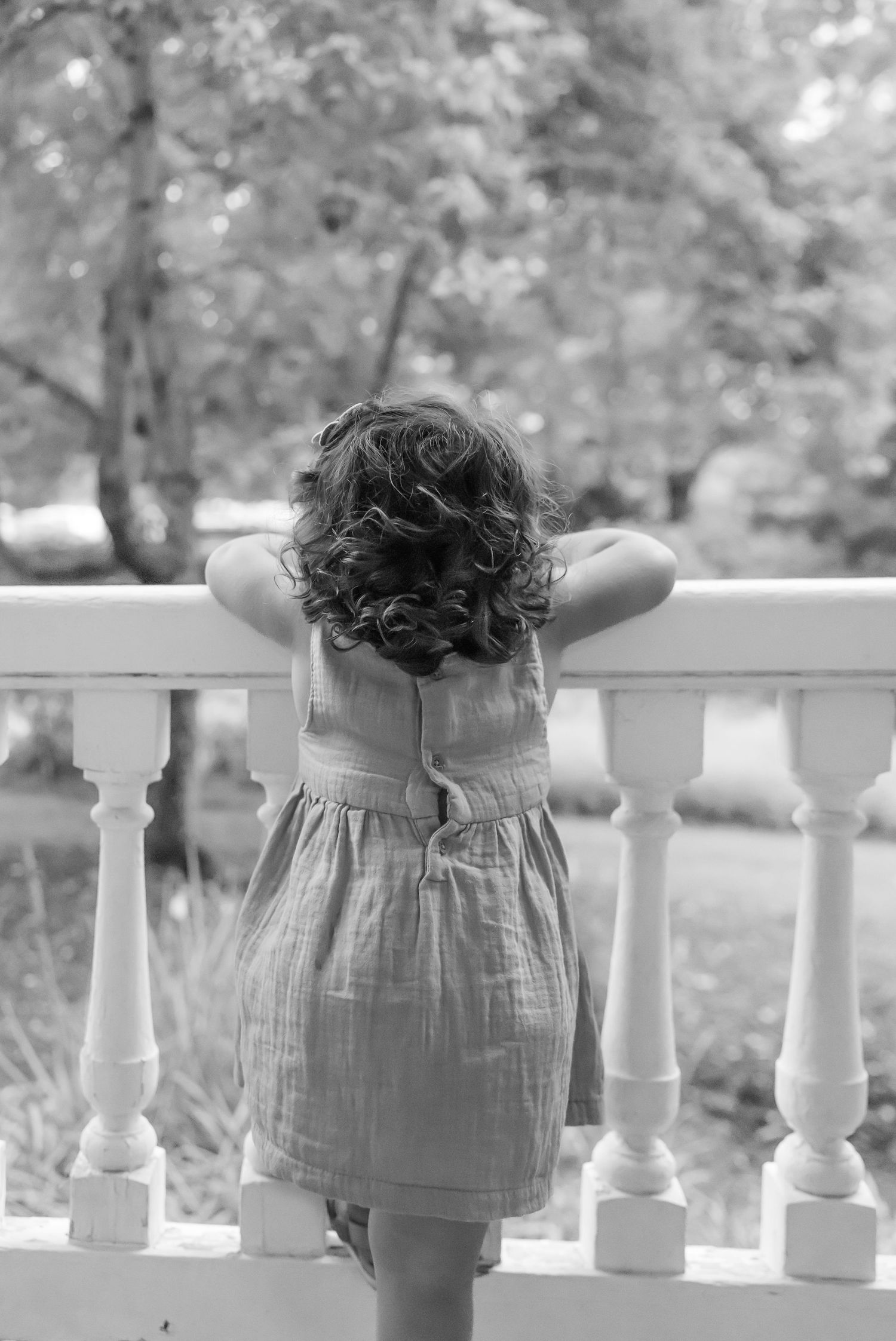 Black and white portrait of a small child in a dress leaning on a white railing, looking out at a garden by Columbus, GA family photographer