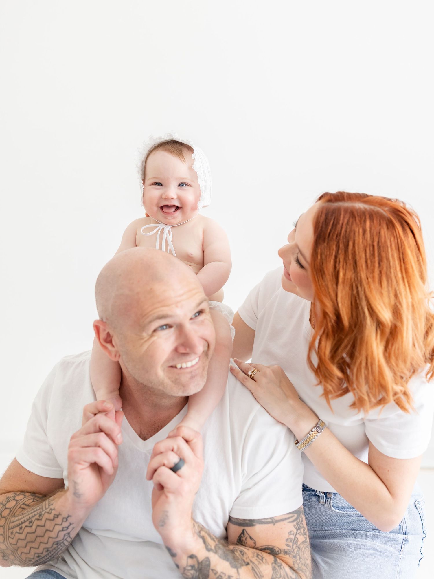 Smiling bald tattooed father holds baby on shoulders while redhead mother looks on, all wearing white.