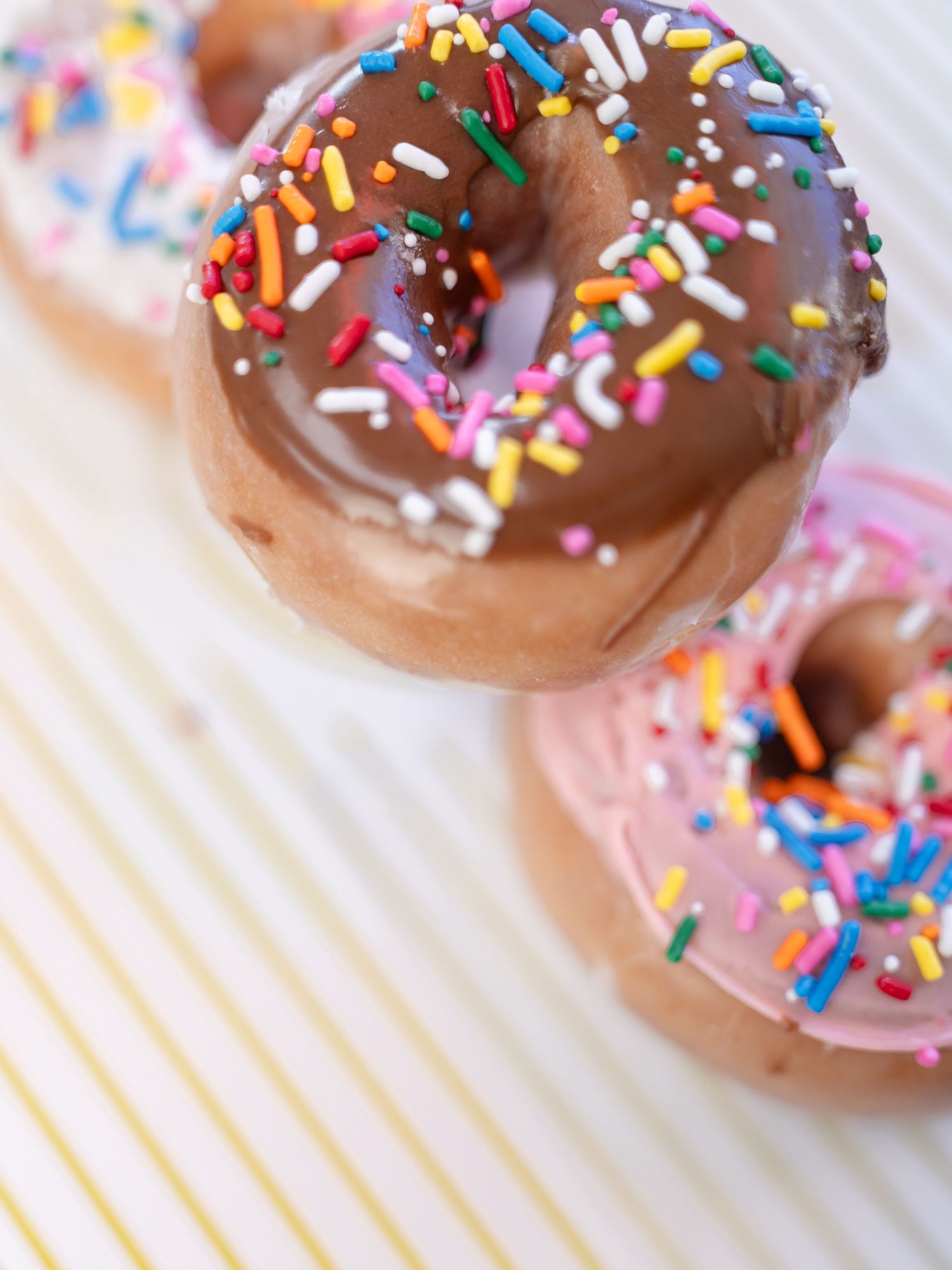 Chocolate glazed donuts with colorful rainbow sprinkles arranged in a row on a light colored surface.