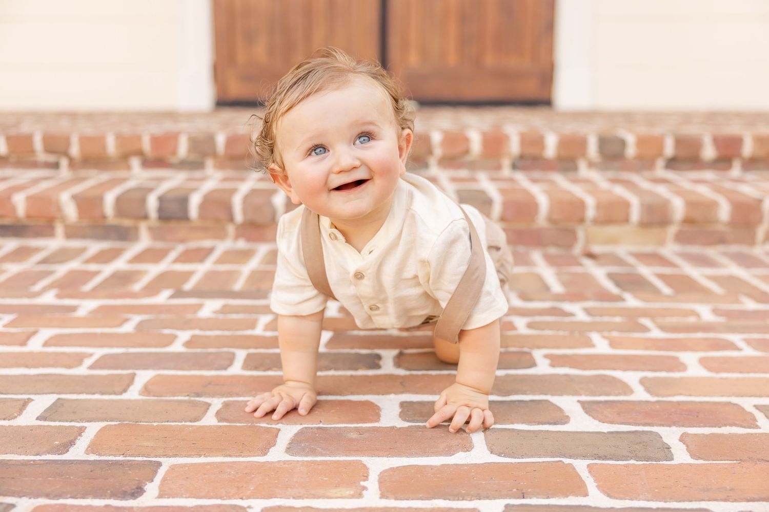 Happy baby in beige romper crawling on brick steps, smiling and looking up outdoors.