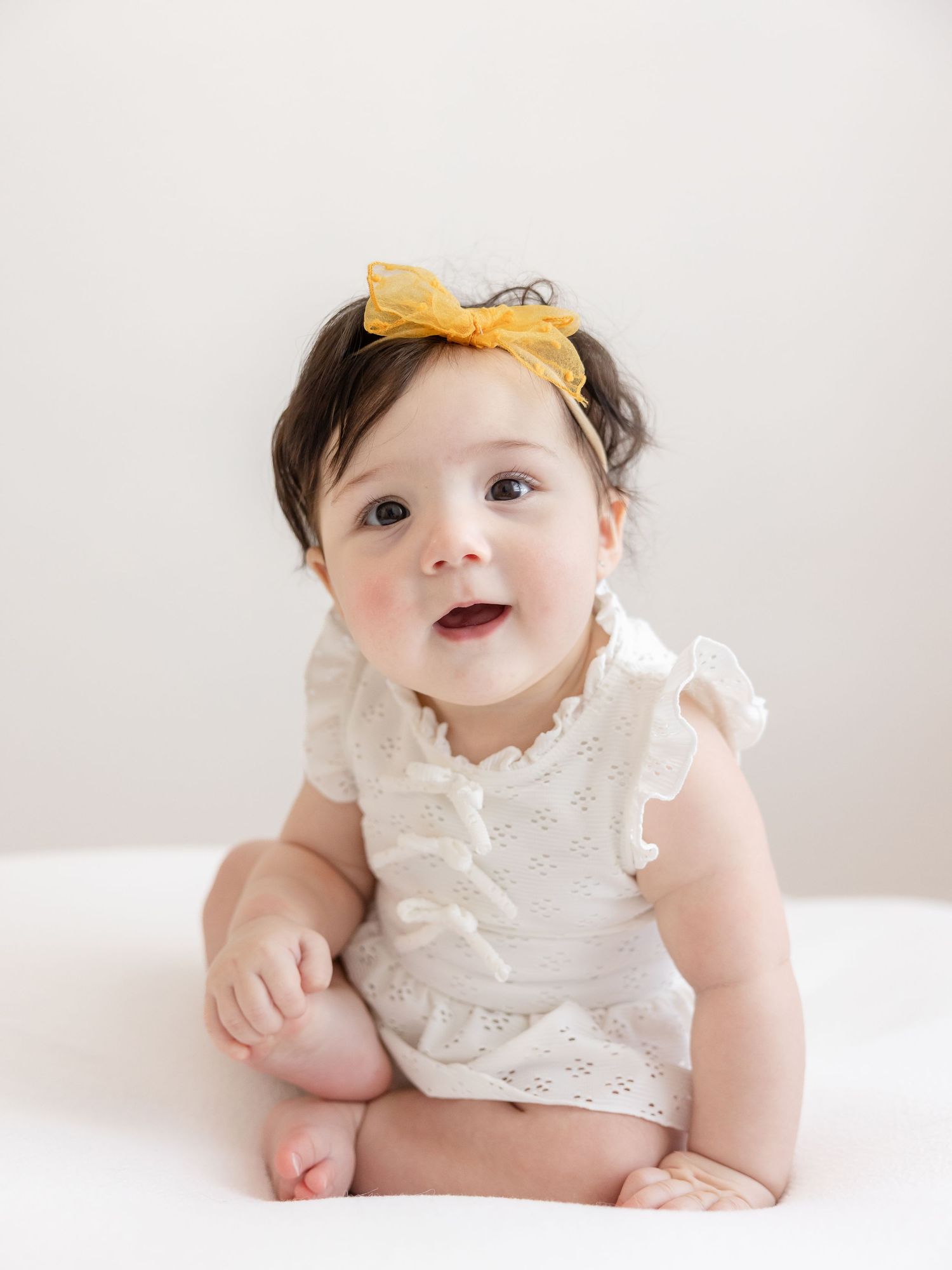 Adorable baby girl in white eyelet romper and yellow bow headband sitting up and smiling at camera.