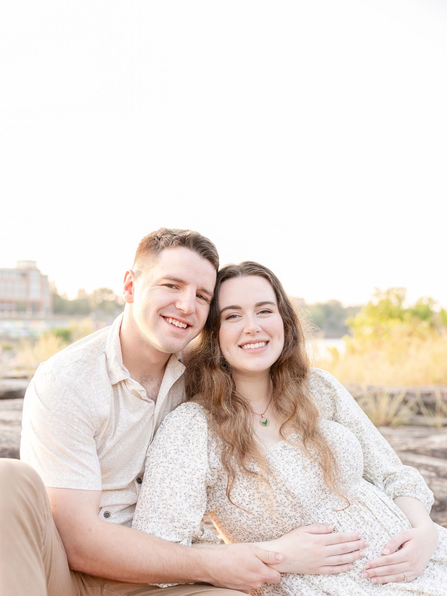 A couple in coordinating white outfits sits together on a sandy beach at sunrise for their Columbus, GA maternity portrait session.