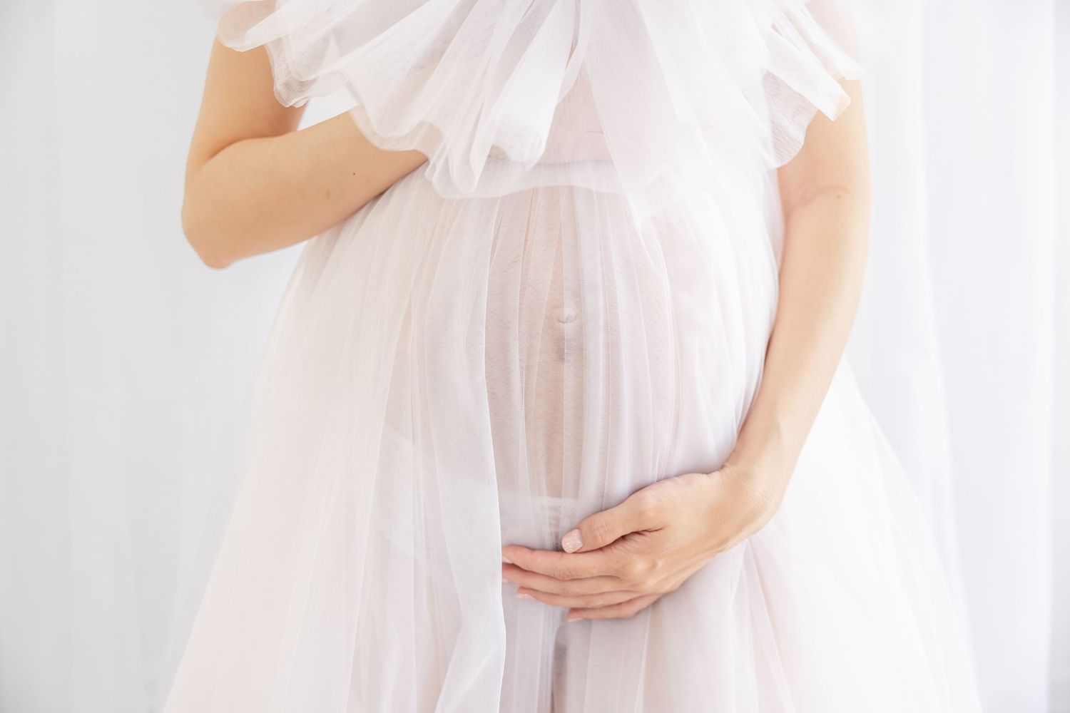 A pregnant woman in a flowing white chiffon dress cradles her baby bump against a light background.