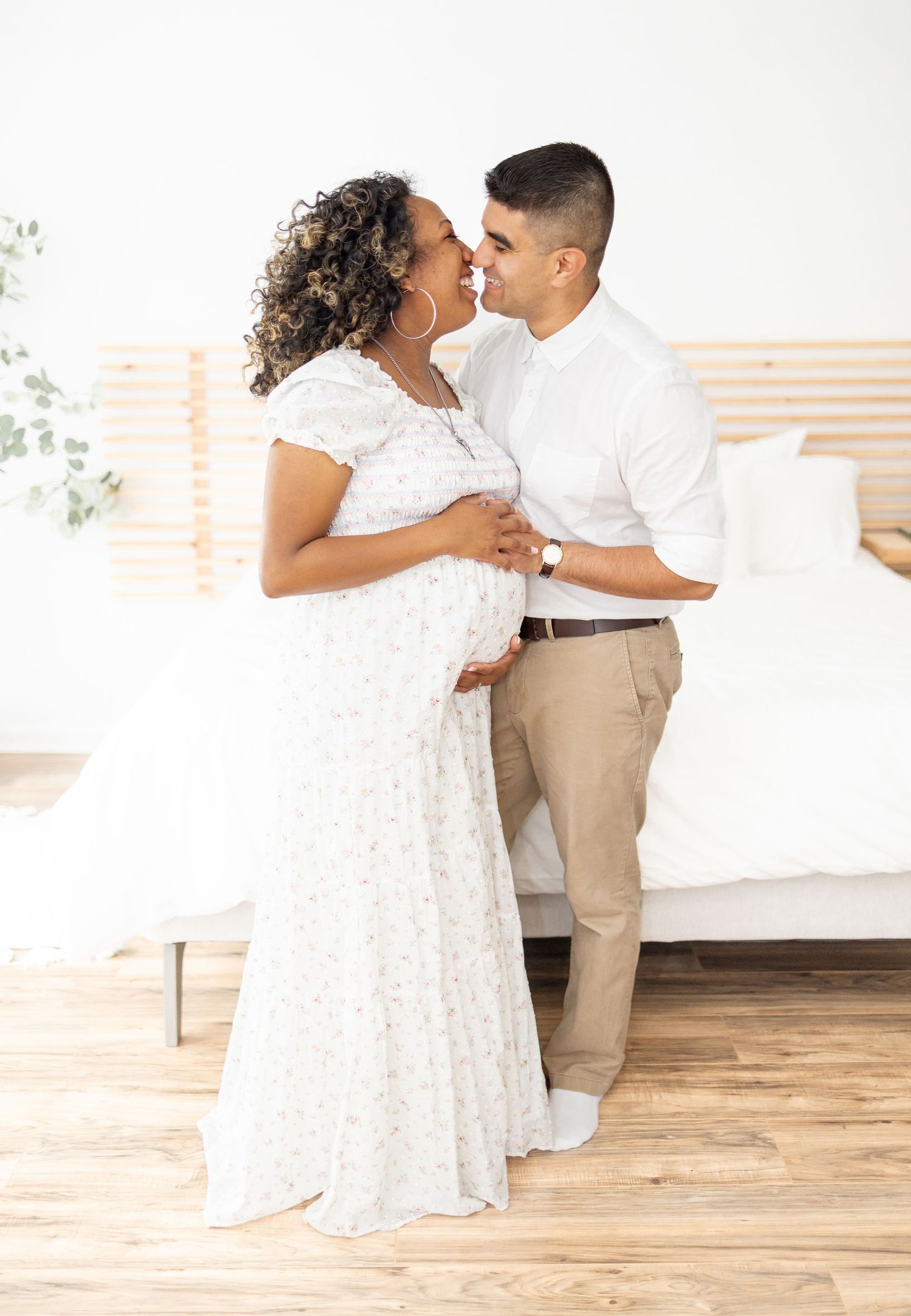 A couple in white attire share an intimate moment in a bright, minimalist bedroom setting with hardwood floors.