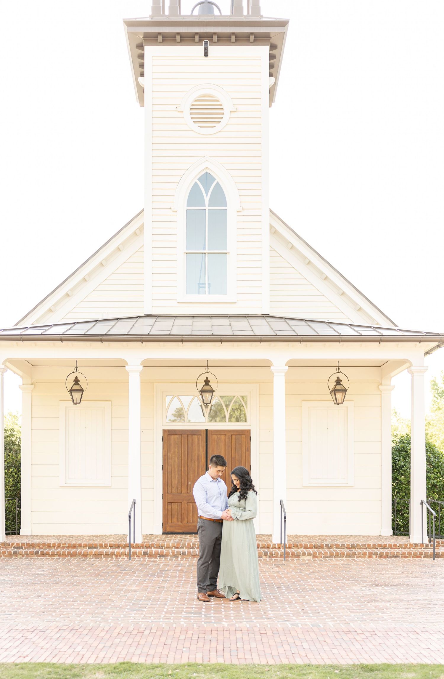 Couple embracing in front of a white chapel with arched window and brick pathway during their maternity portraits in Columbus, GA.