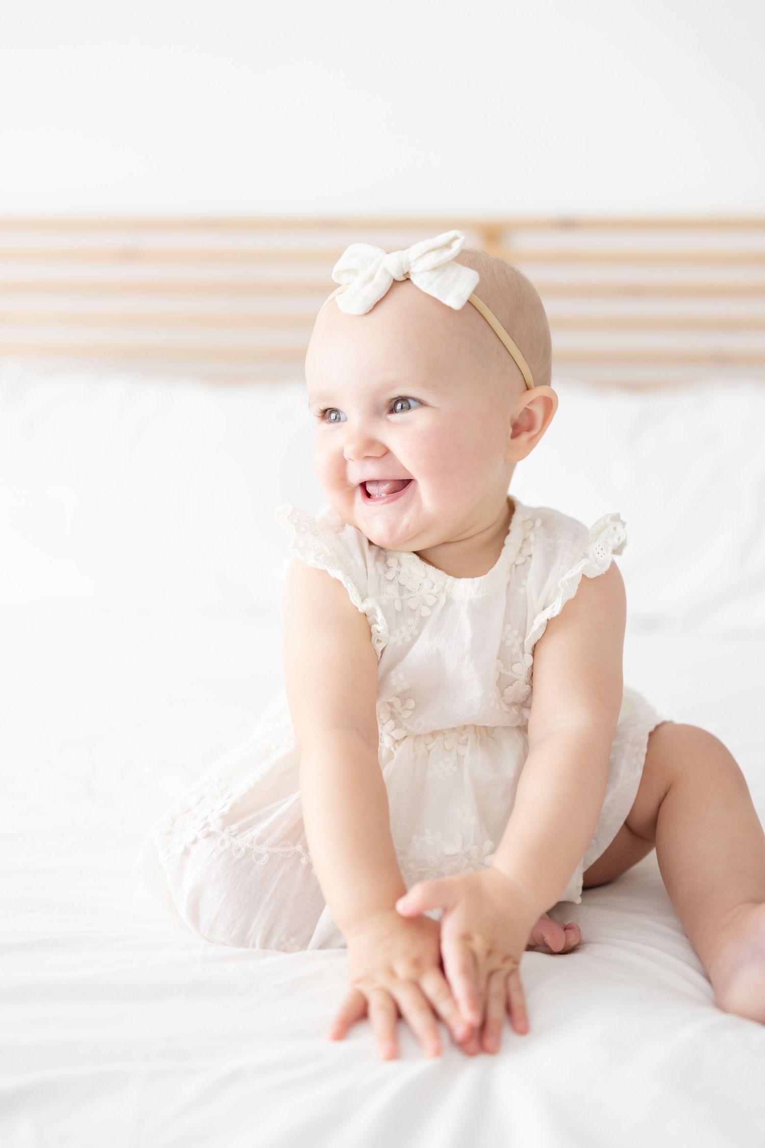 Smiling baby girl in white lace romper and bow headband sitting on white bed.