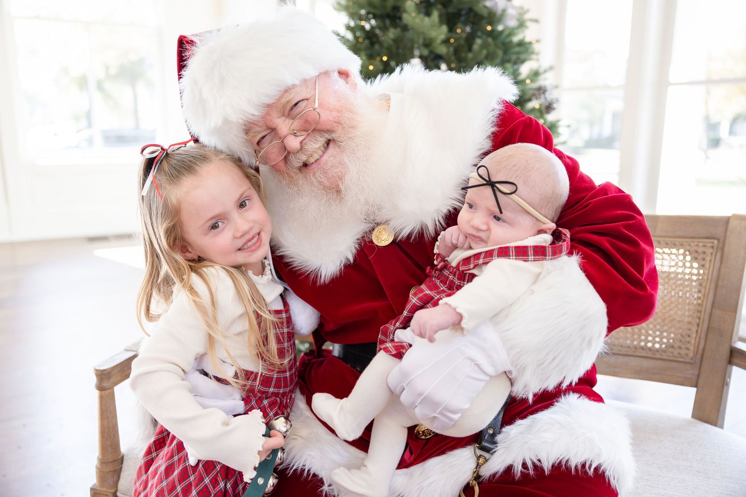 Two children sitting on Santa Claus's lap smiling for a Christmas photo near a decorated tree.