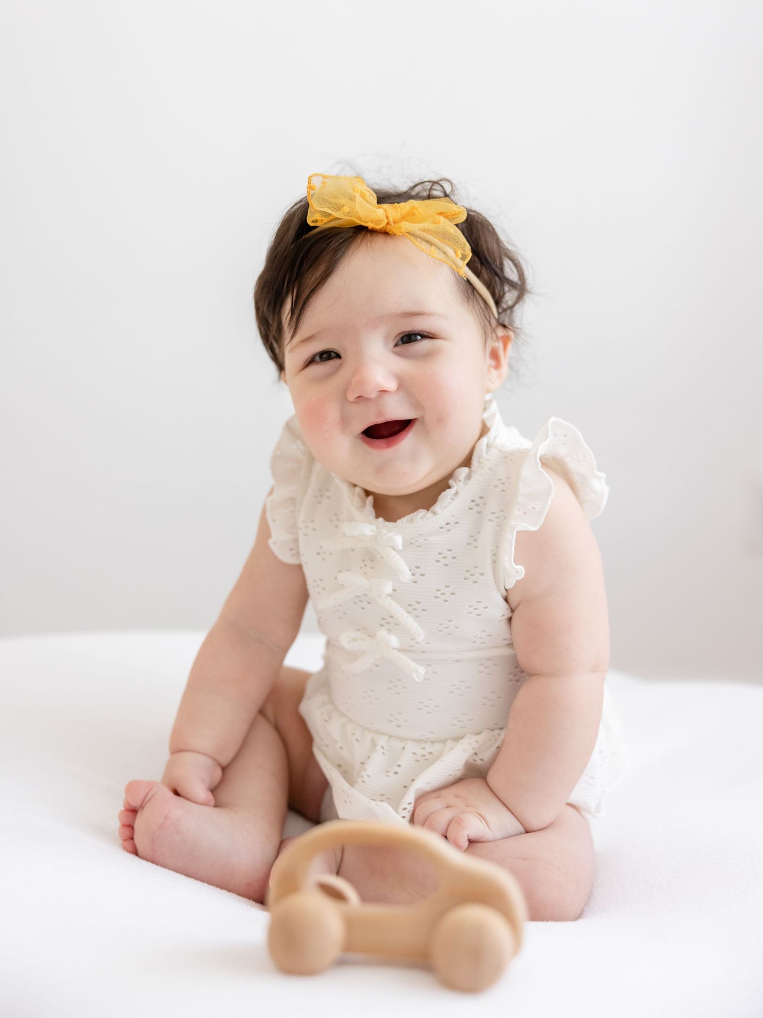 Six month baby girl wearing white lace dress and yellow headband smiles while sitting and smiling during her milestone portrait session.