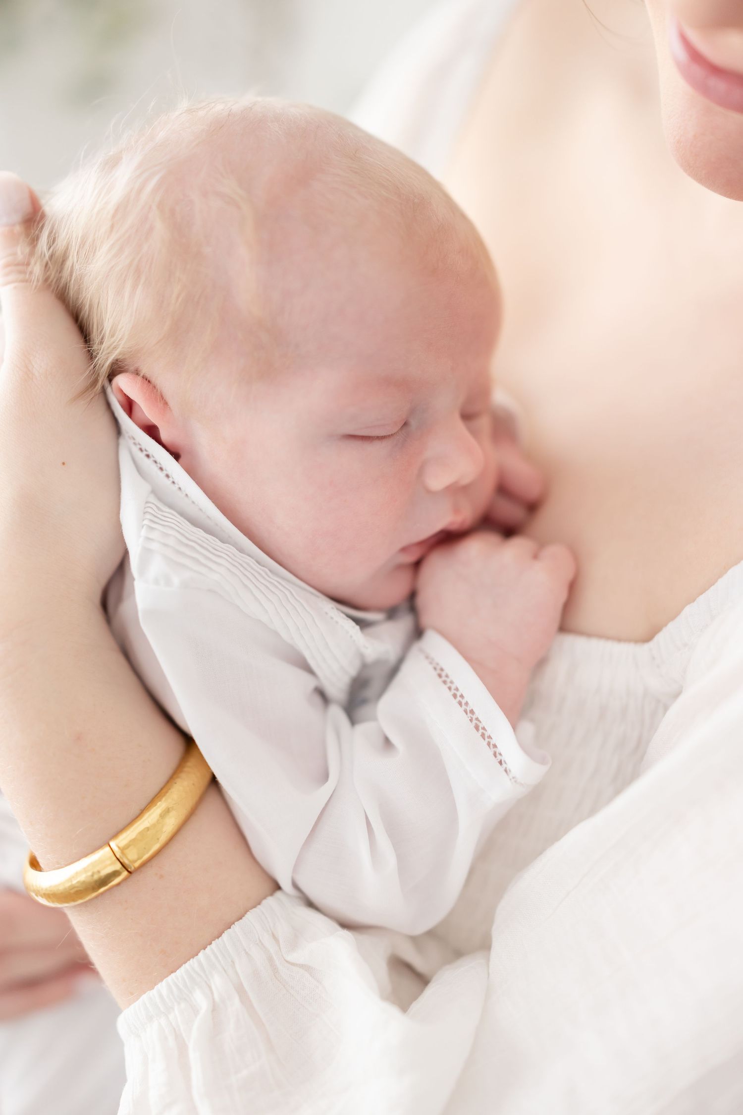 A newborn baby in white newborn gown sleeps quietly on his mother’s chest at his Columbus, GA newborn portrait session.