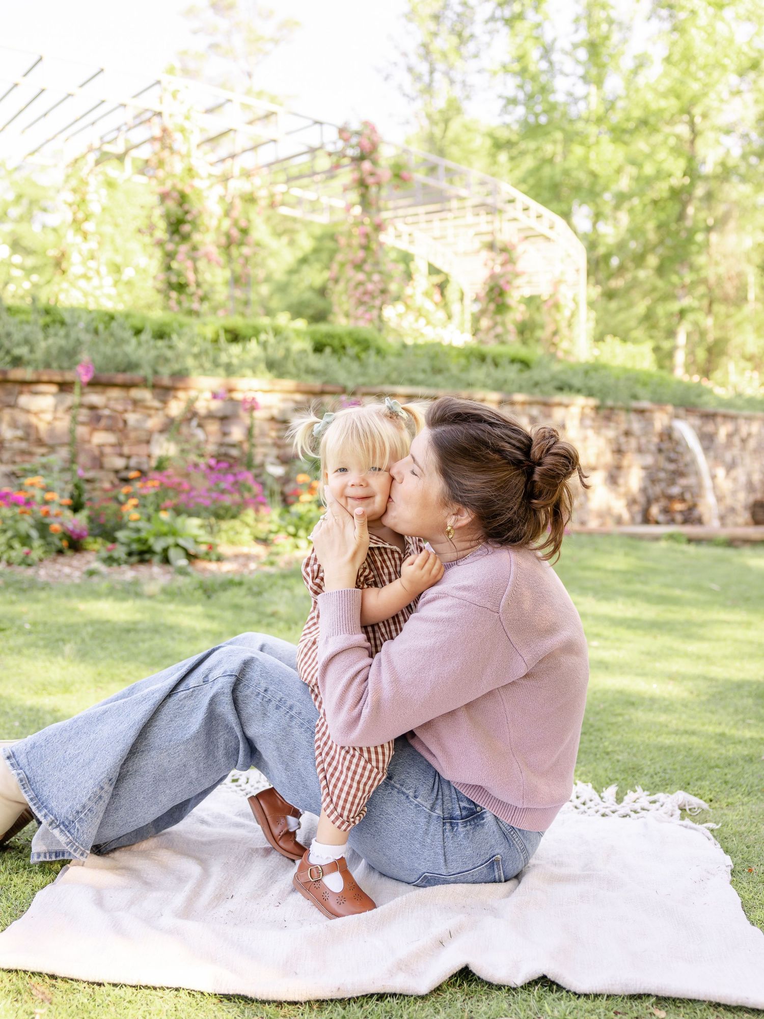 A mother kisses her toddler on a white blanket in a lush garden setting, both wearing pink and denim outfits.