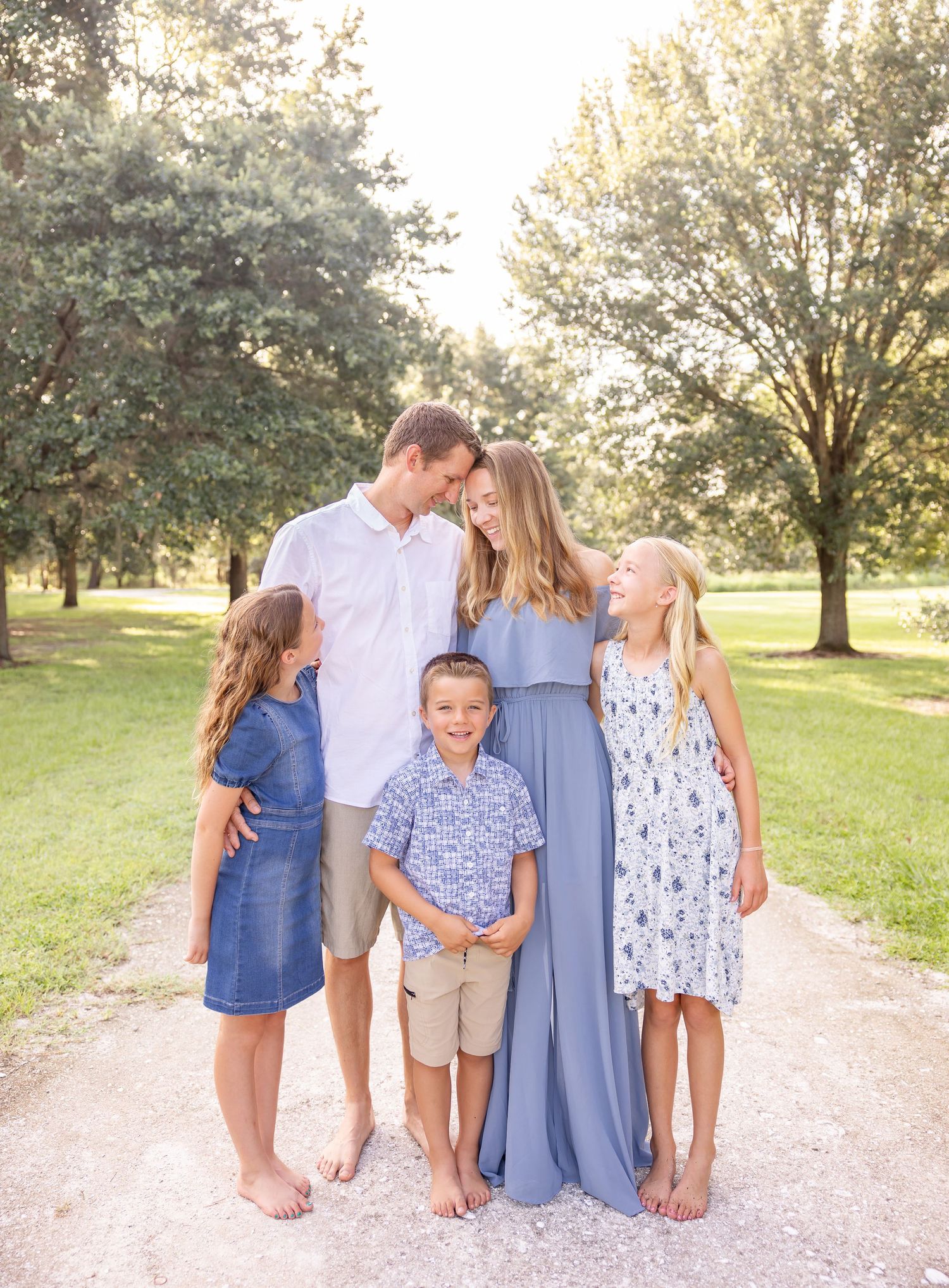 Outdoor family portrait of a group in coordinating blue and white outfits standing together on a gravel path in a sunlit park.