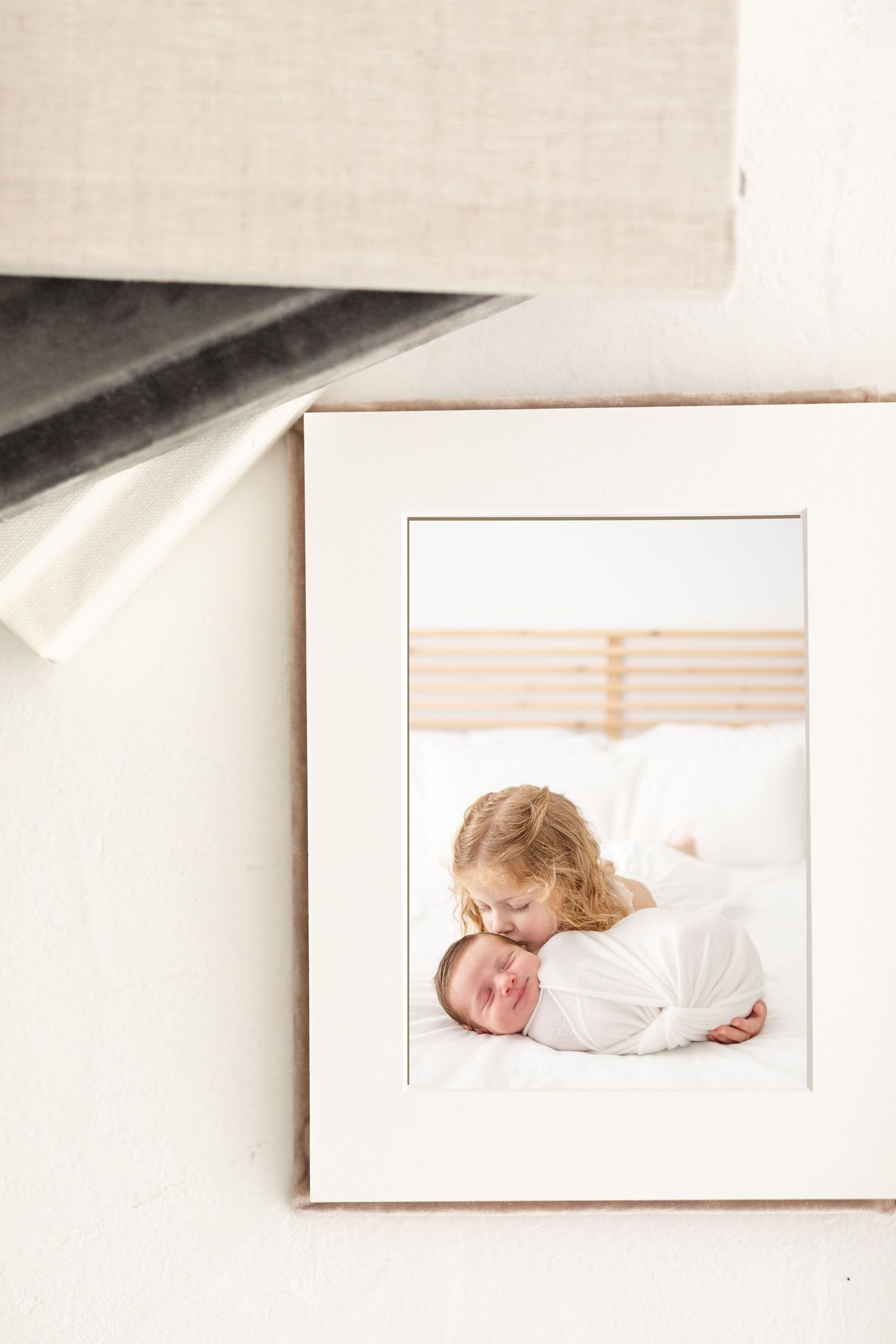Framed photo of a young child kissing a swaddled newborn baby on a white bed, displayed against a wall.