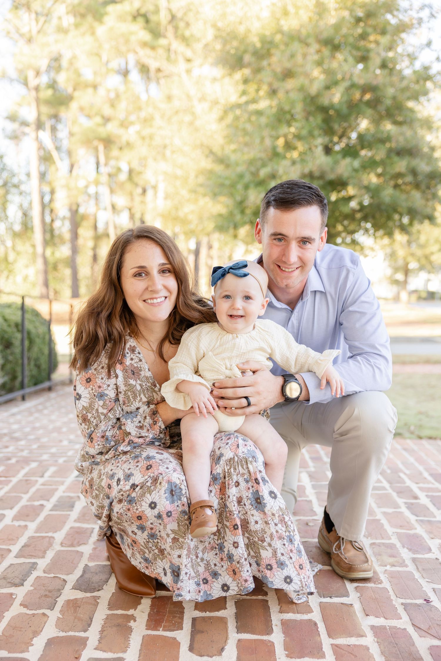 Happy young family with baby girl in floral dress posing outdoors on brick path surrounded by trees.