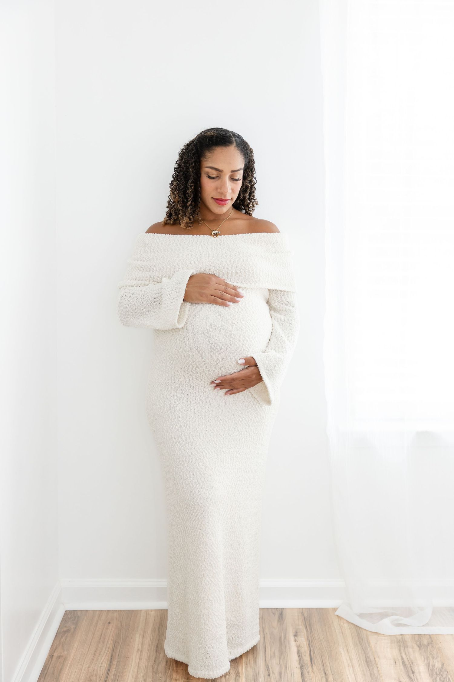 Pregnant woman in white off-shoulder knit dress cradling baby bump, standing against white wall.