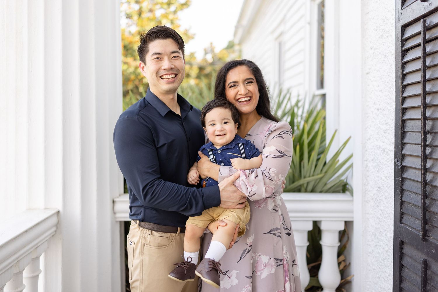 Happy Asian family with parents holding baby boy smiling on white porch of home.
