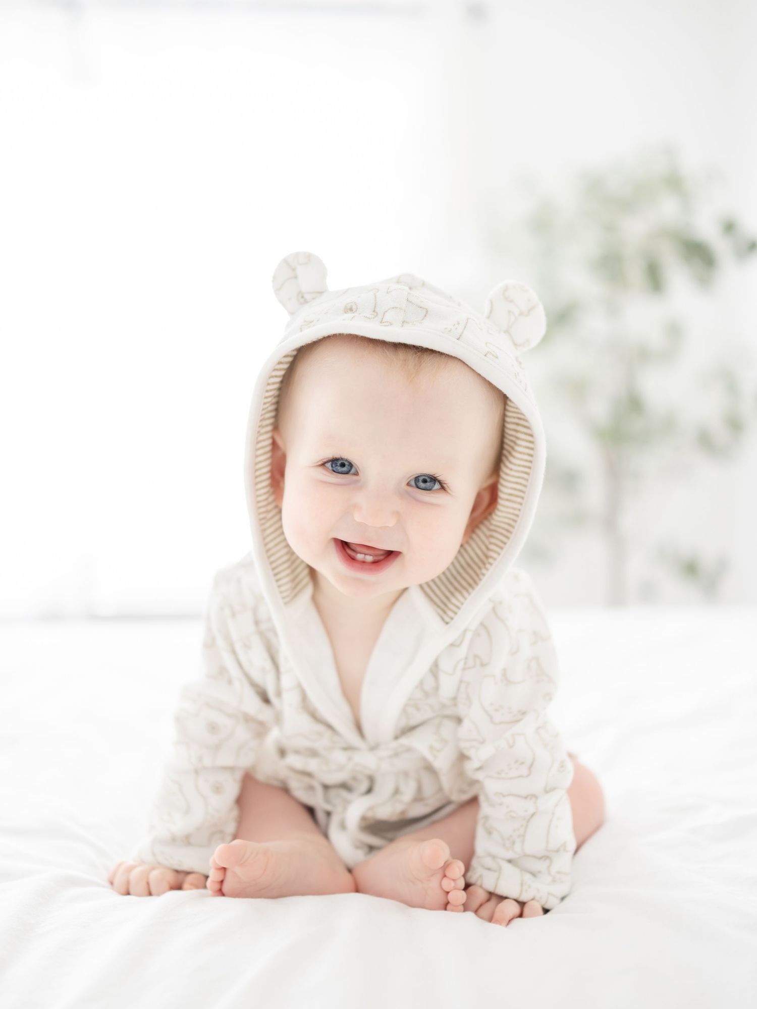 A baby wearing a baby bath robe with little bear ears sits on white bedding in a bright, airy setting at his Columbus, GA milestone portrait
