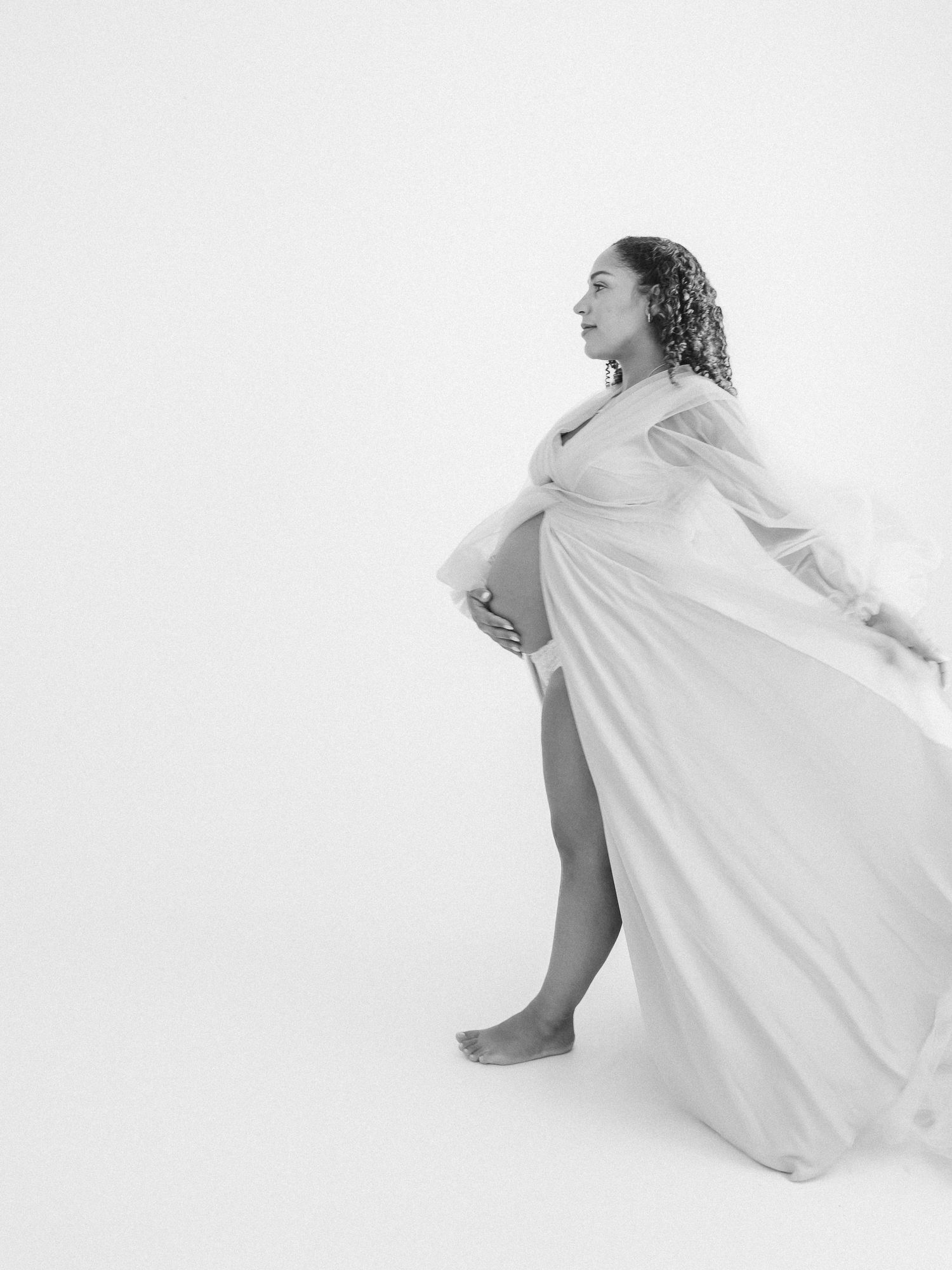 Black and white maternity photo of a pregnant woman in flowing white gown, gazing sideways in a bright studio.