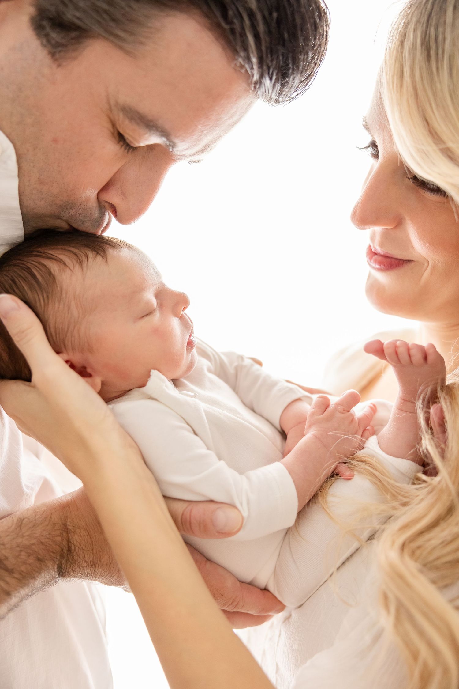 Parents tenderly holding and kissing their sleeping newborn baby dressed in white, sharing a loving family moment.