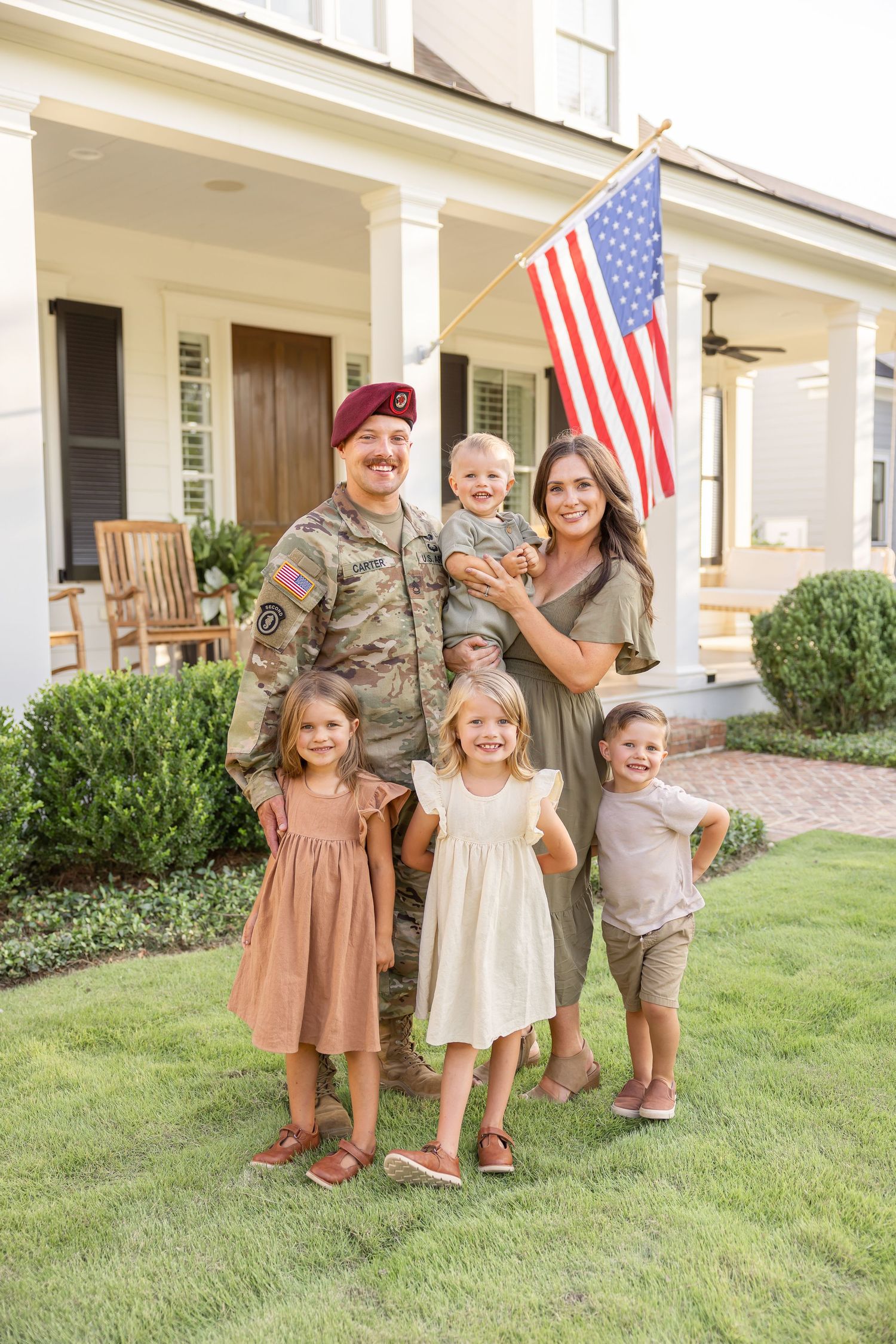 A military servicemember stands with family in front of a classic white porch with American flag.