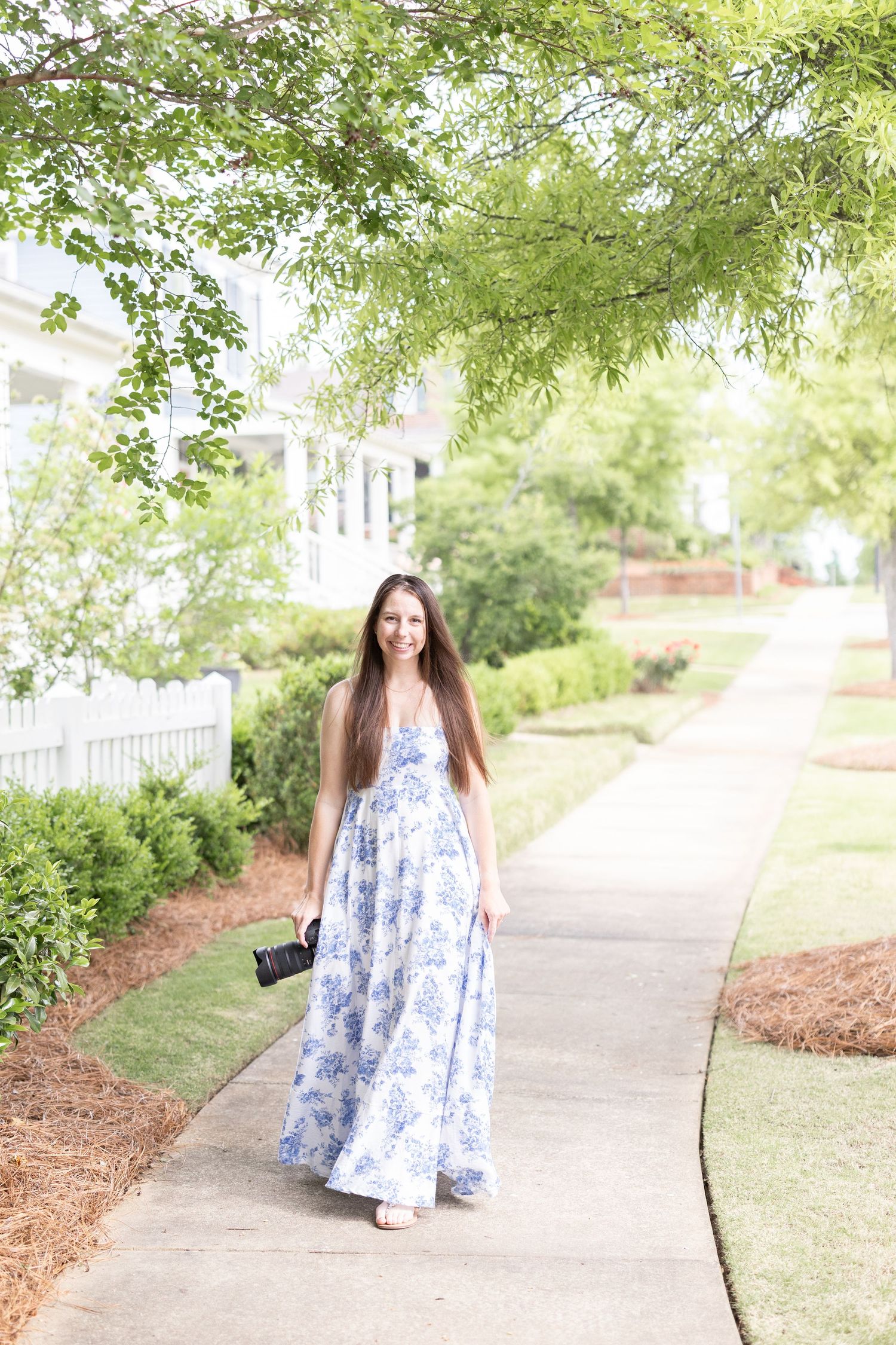 A woman in a flowing blue and white floral maxi dress walks along a curved garden pathway on a sunny day.