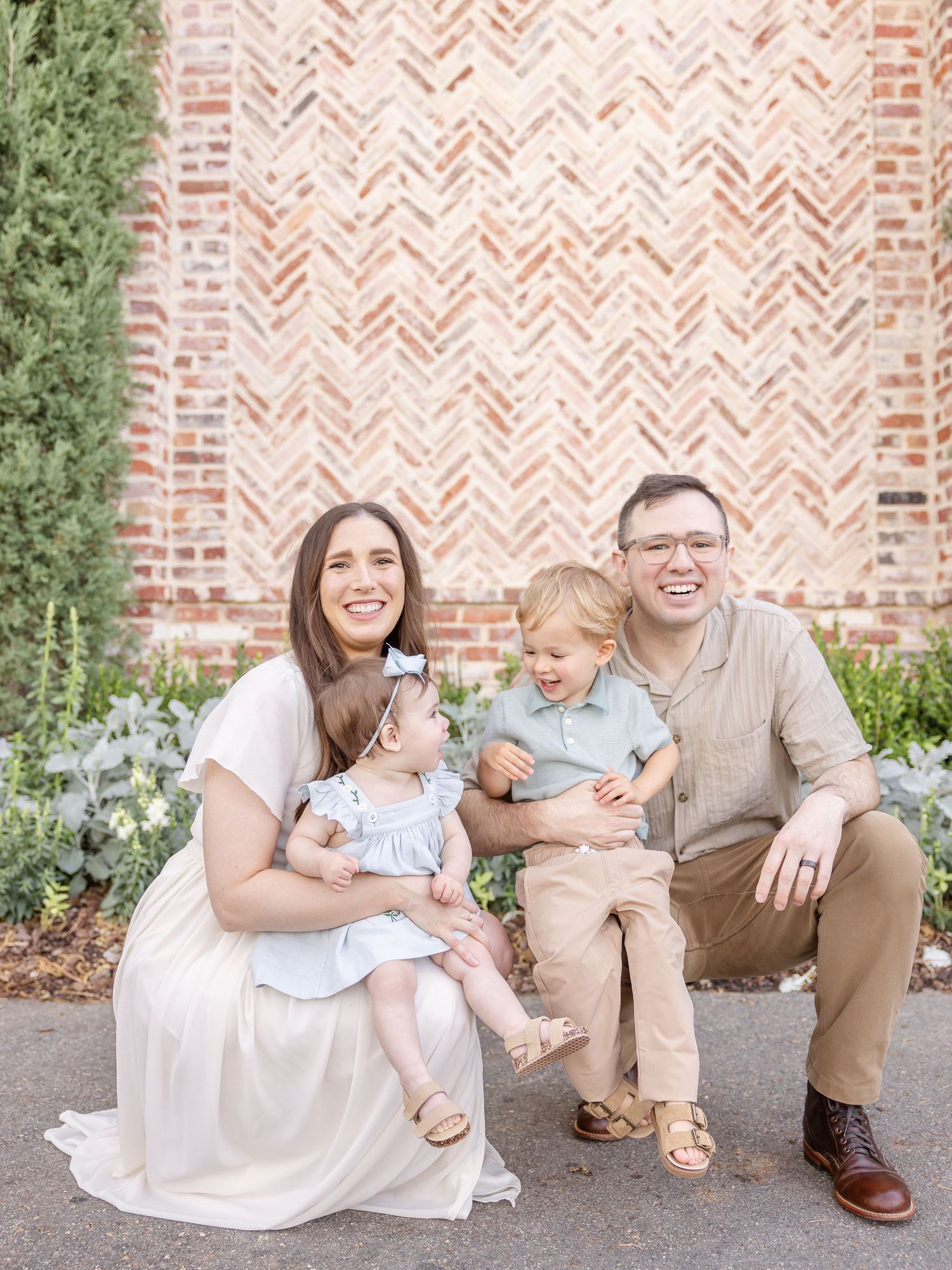 Happy family of four posing in front of a herringbone brick wall, parents smiling with two young children outdoors.
