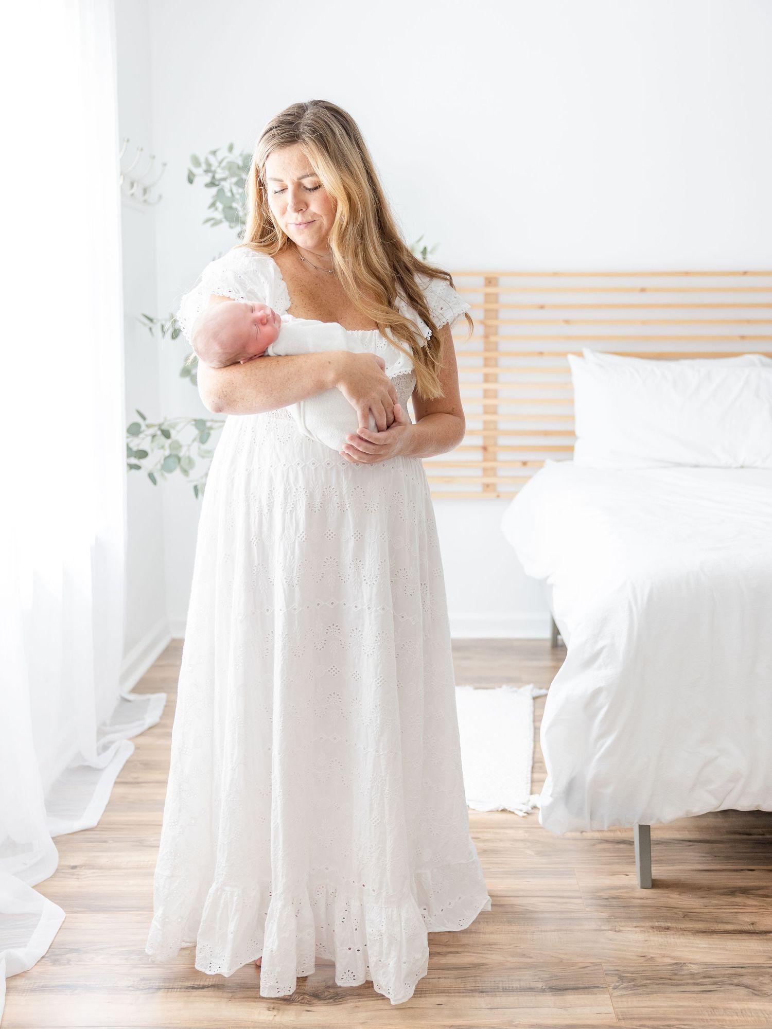 A mother in a white lace dress tenderly holds her newborn baby in a bright, airy bedroom.