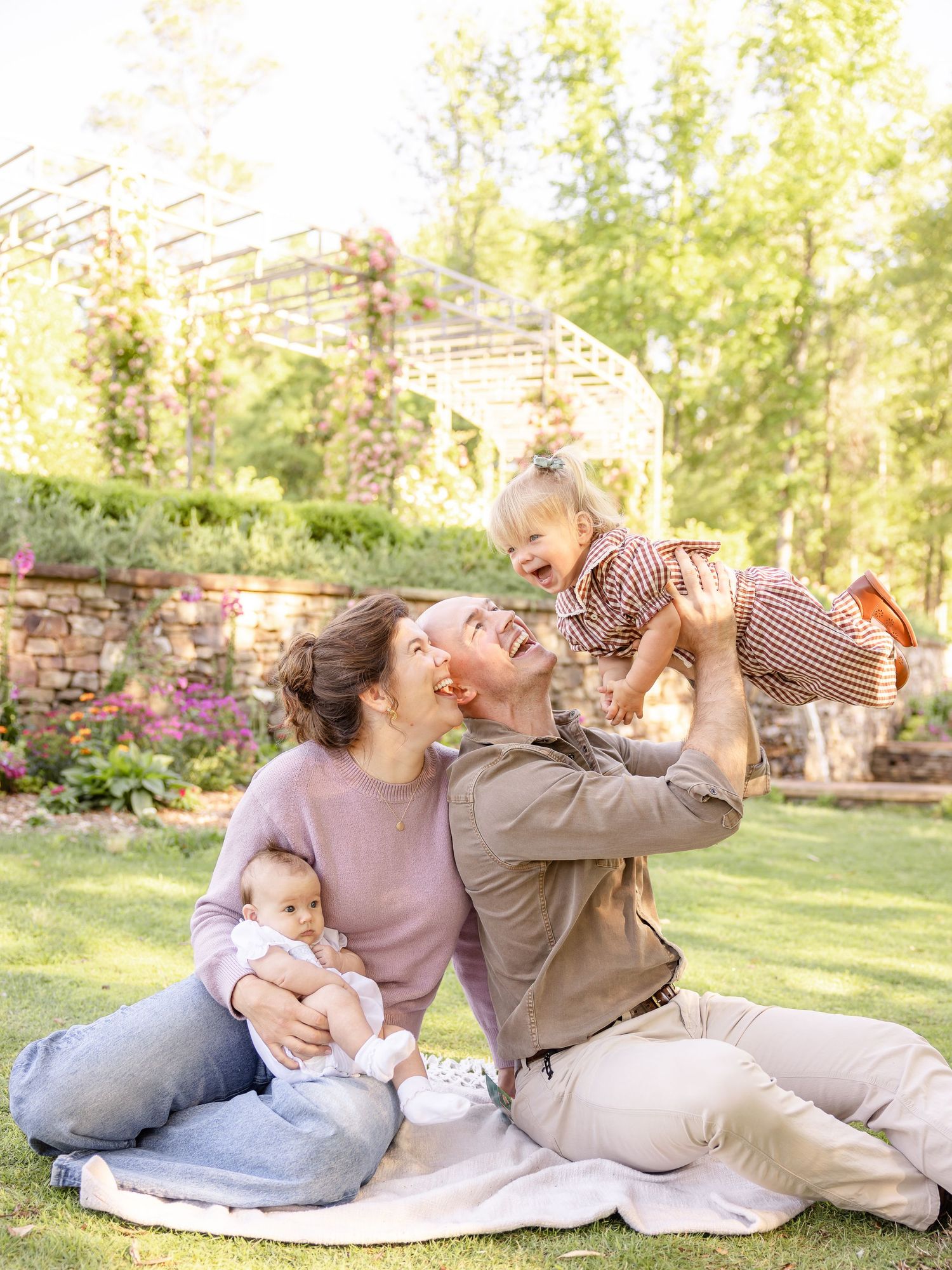 Happy family sitting on grass in a garden, parents laughing with two young babies on a sunny day.